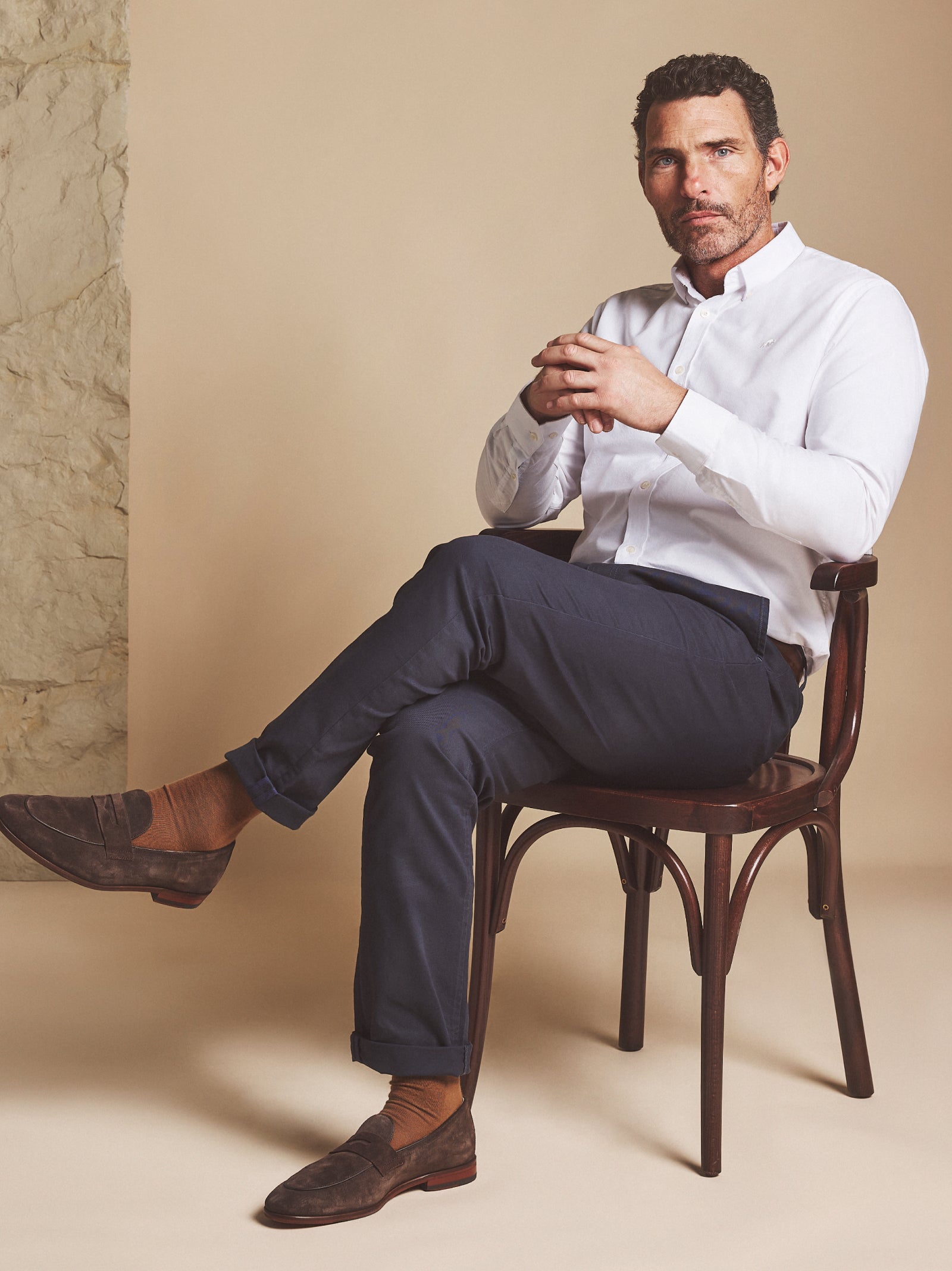 A man wearing navy chino trousers and a white shirt in front of a beige coloured wall. 