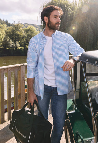 Man in a light check shirt and jeans standing by a car near a river, holding a duffel bag.