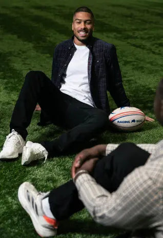 Man in casual Raging Bull outfit sitting on indoor turf with a rugby ball, chatting with another person.