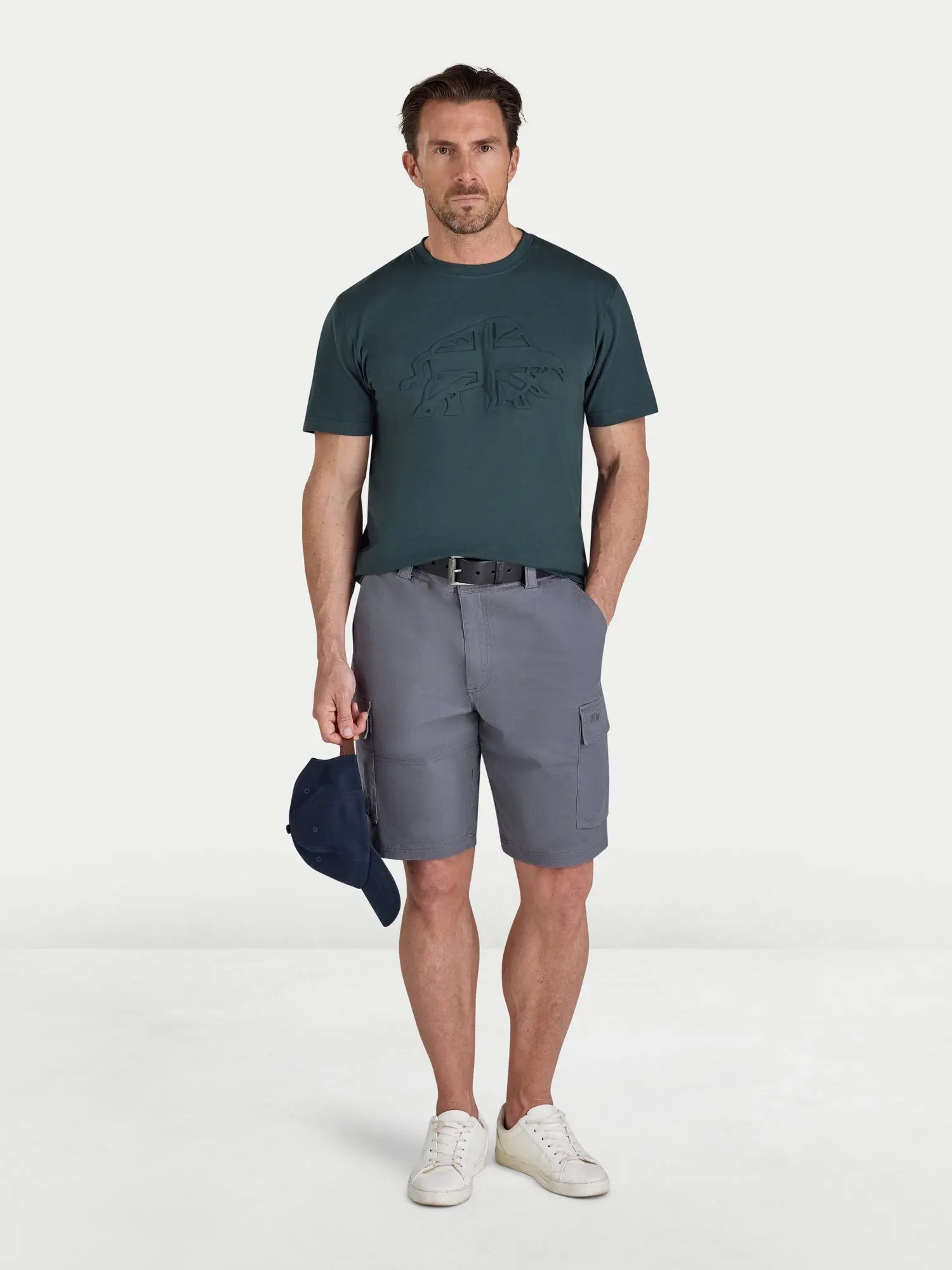Man wearing a green t-shirt and gray shorts holding a cap on a white background