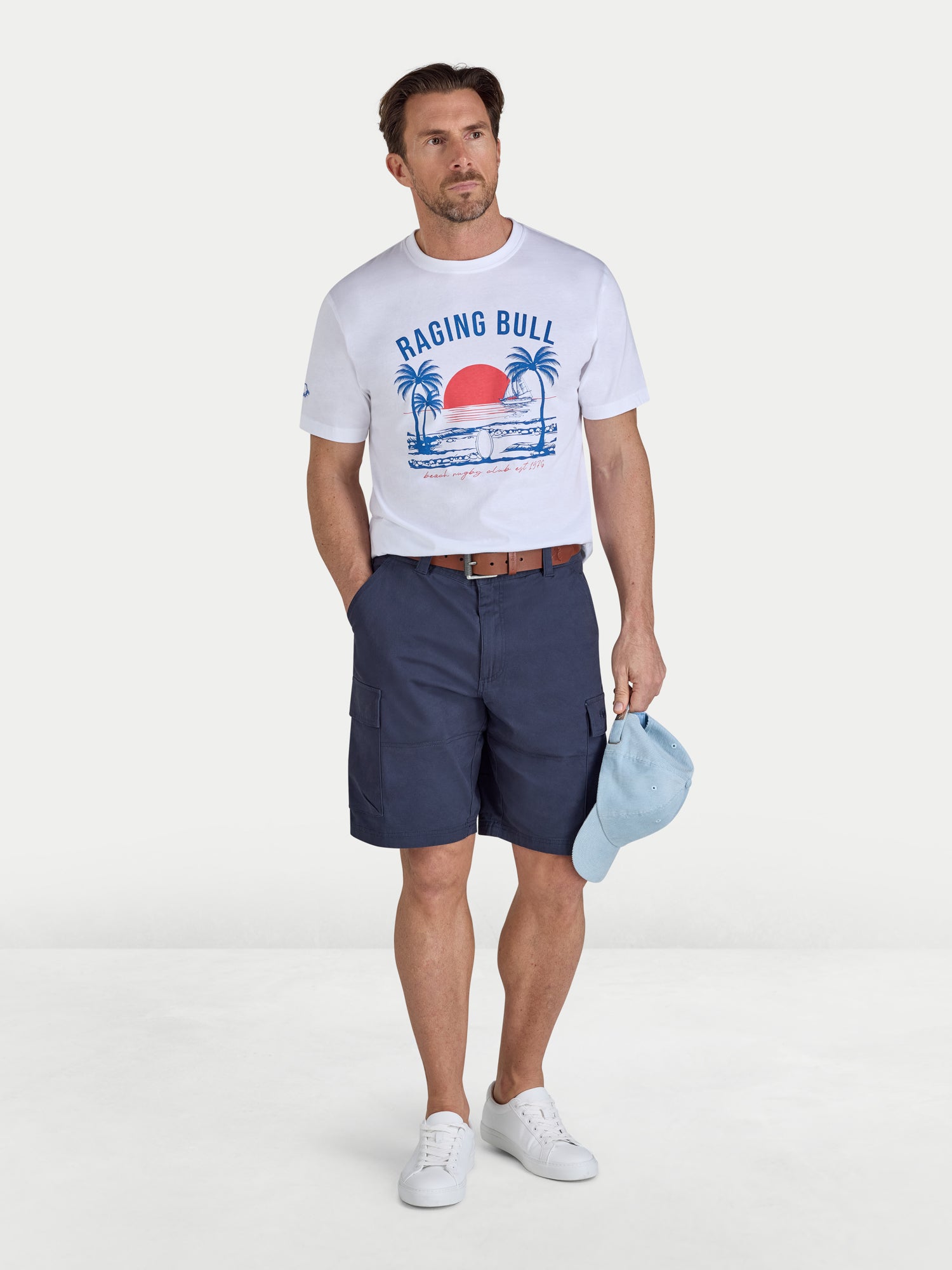 Man wearing a 'Raging Bull' t-shirt, navy shorts, and white sneakers on a white background