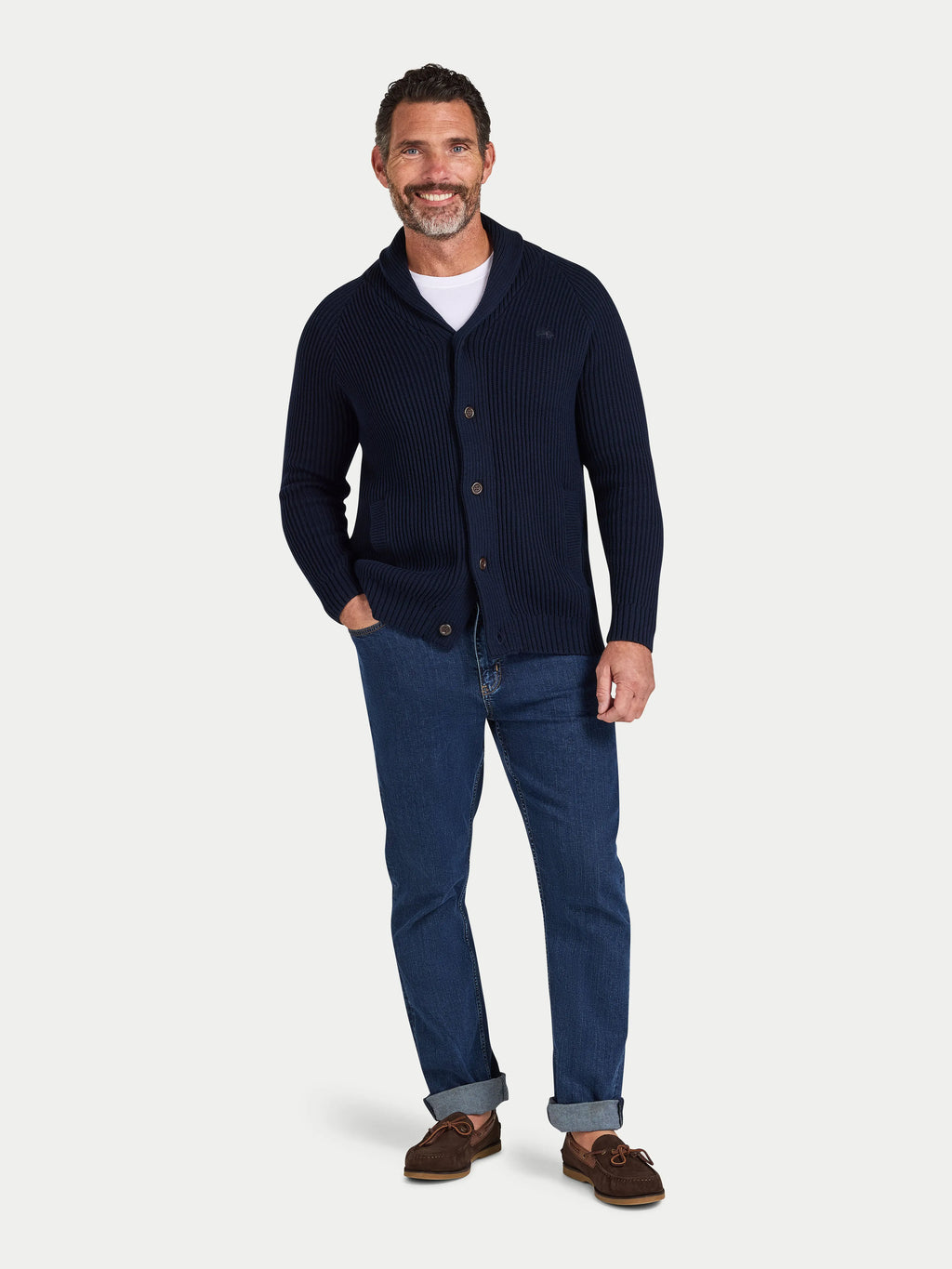 Man wearing a navy cardigan, white shirt, blue jeans, and brown shoes on a light gray background