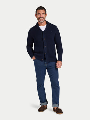 Man wearing a navy cardigan, white shirt, blue jeans, and brown shoes on a light gray background