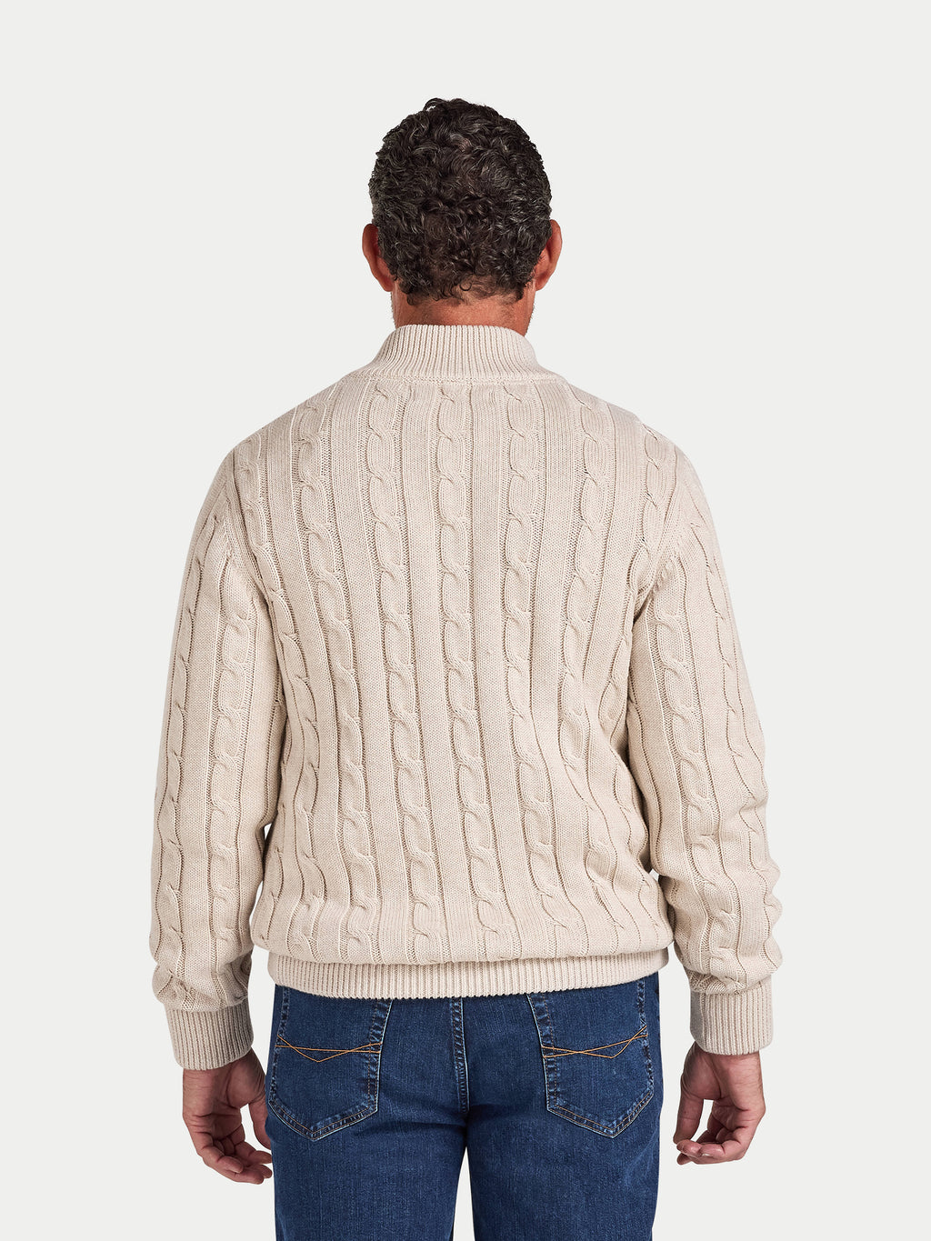 Man wearing a cream cable knit sweater and blue jeans on a white background