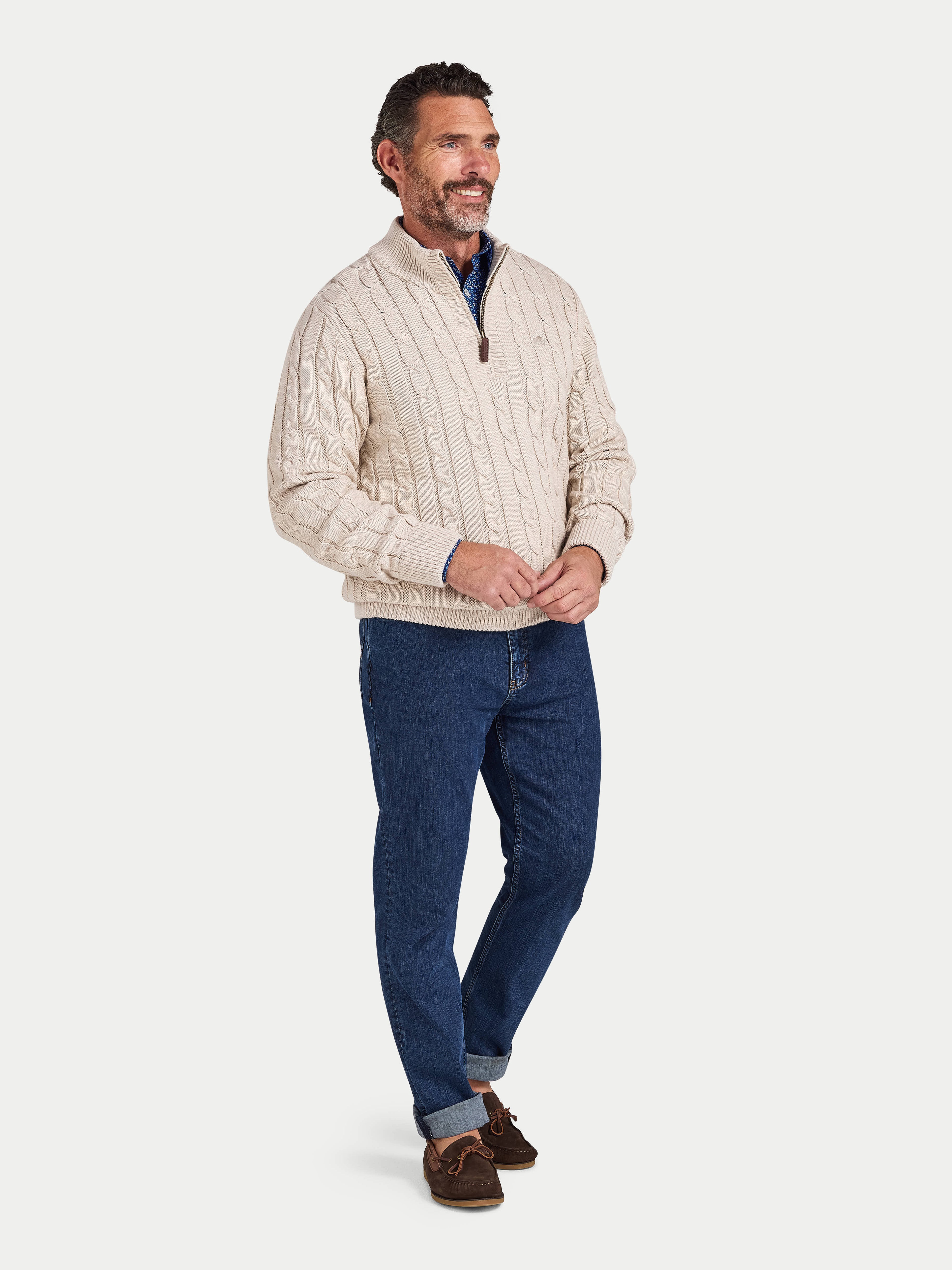 Man wearing a beige cable knit sweater and blue jeans on a white background