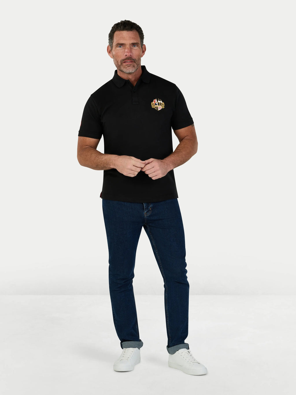 Man wearing a black polo shirt with a logo and blue jeans on a white background