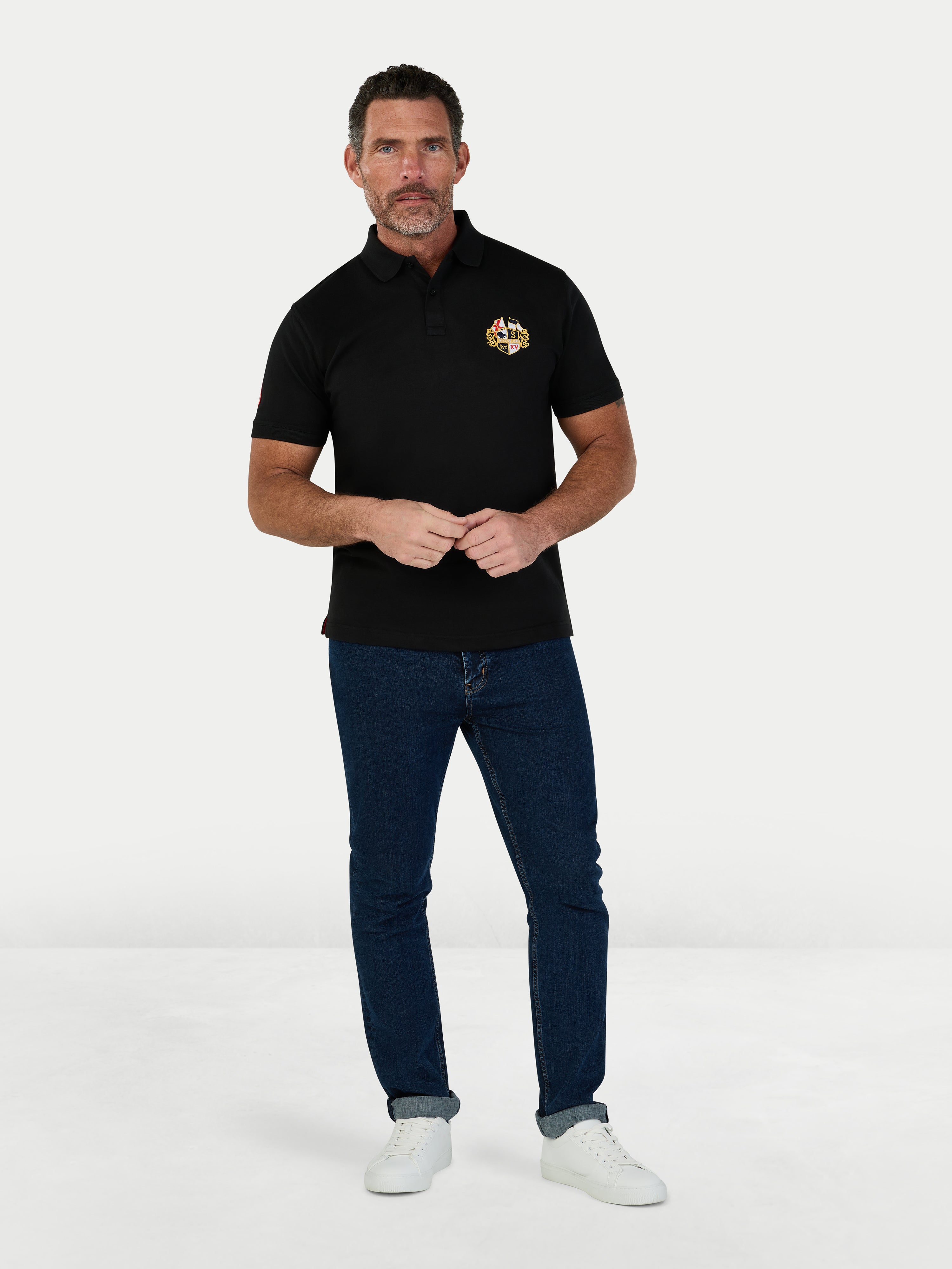 Man wearing a black polo shirt with a logo and blue jeans on a white background