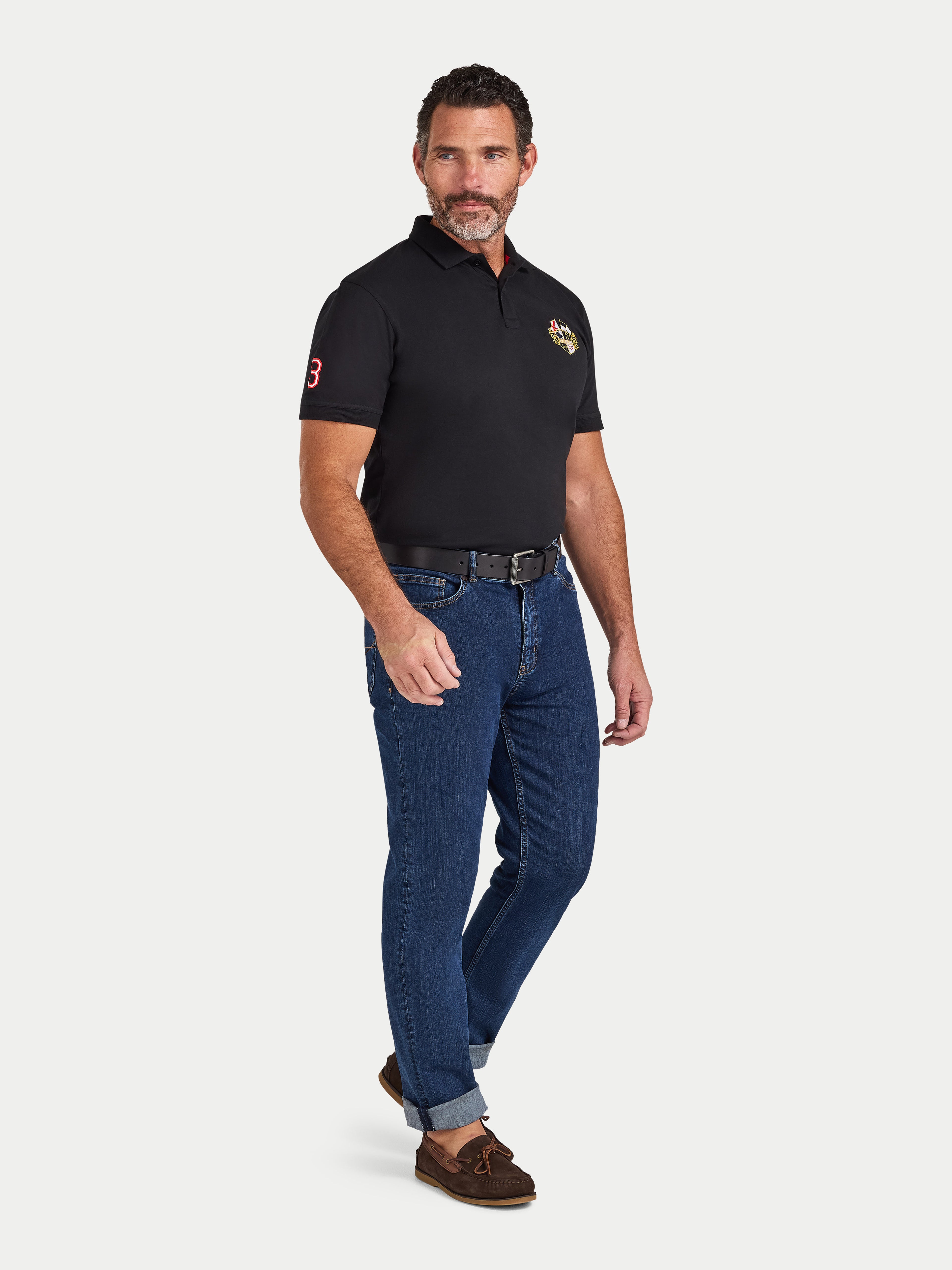 Man wearing a black polo shirt with a logo and blue jeans on a white background