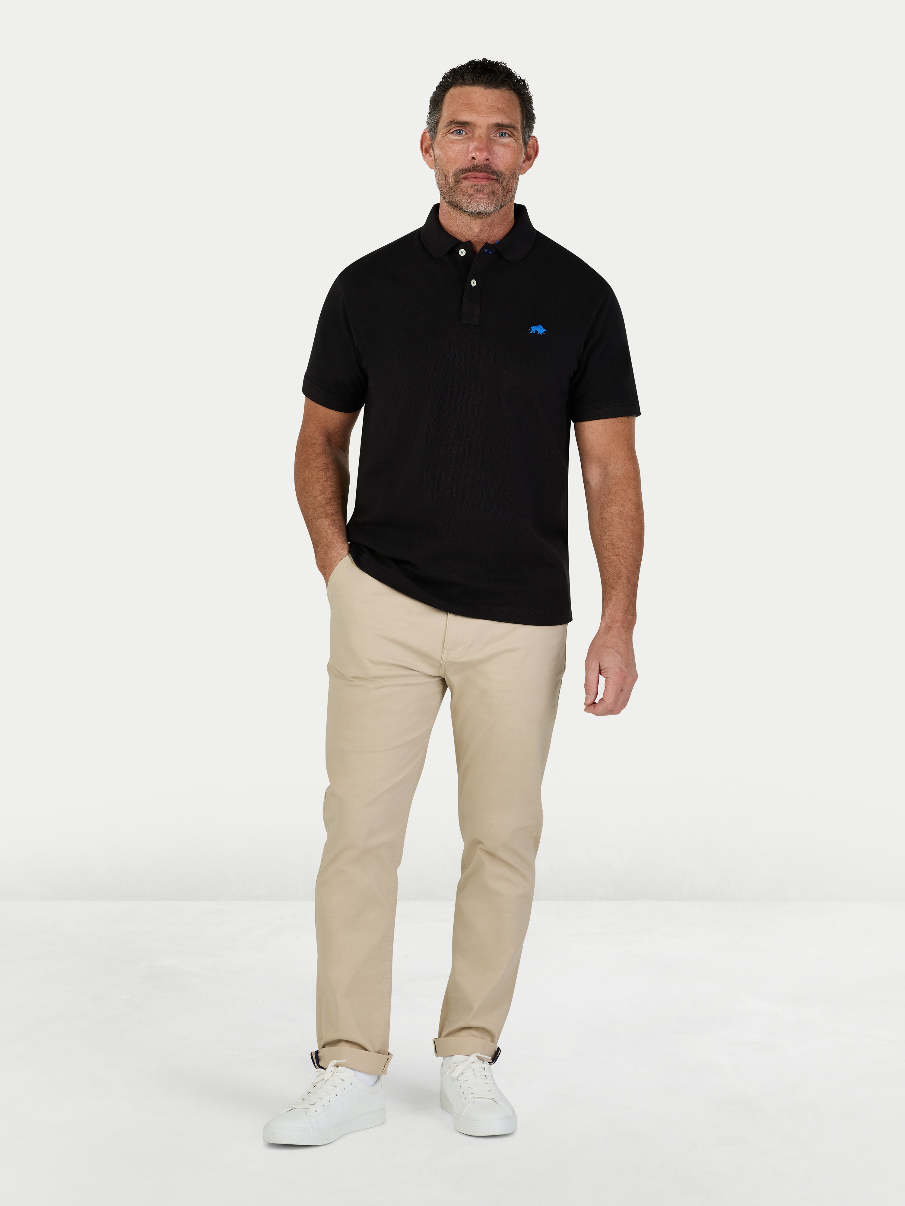 Man wearing a black polo shirt with a logo and beige pants on a white background.