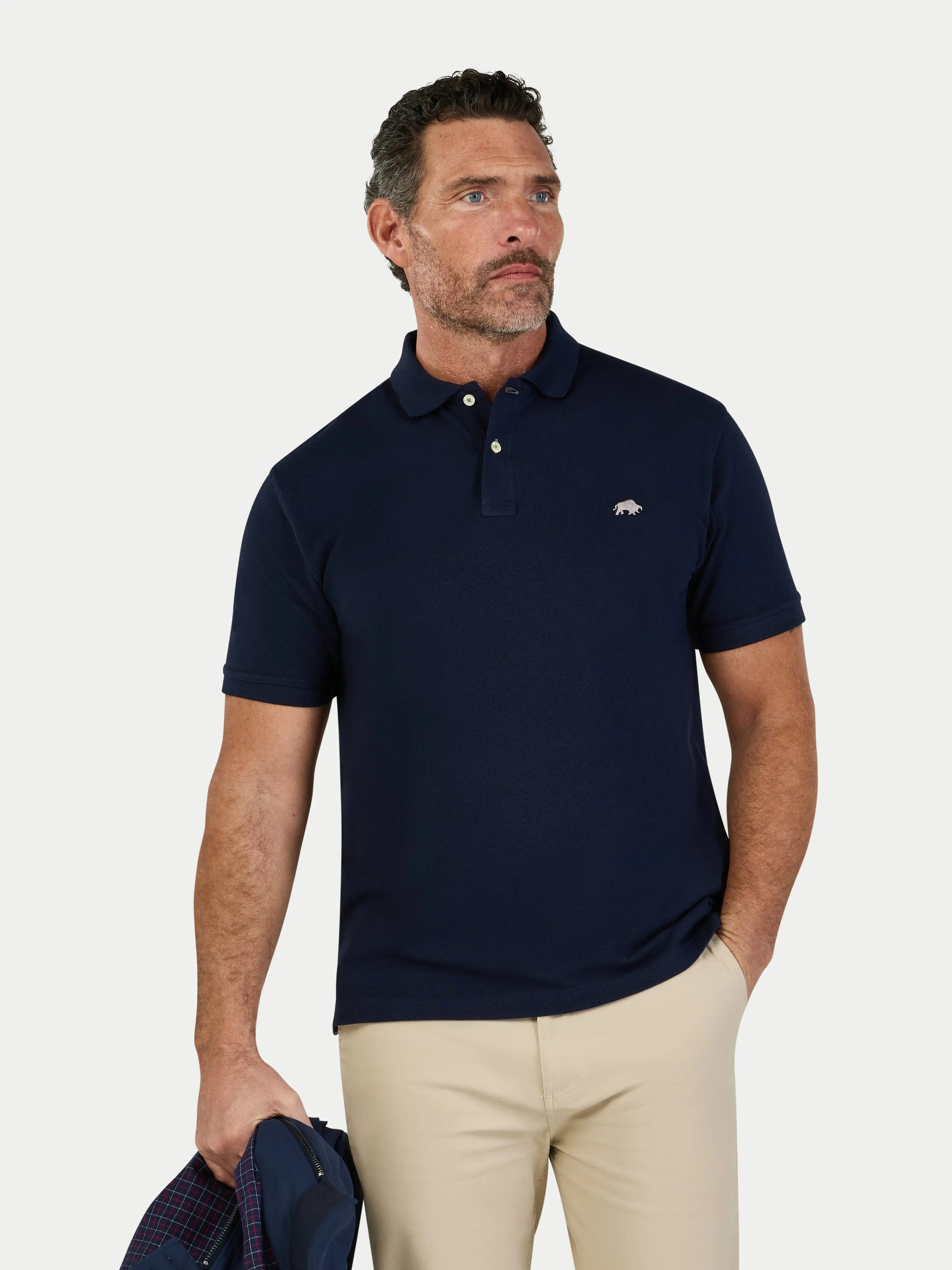 Man wearing a navy polo shirt with a logo, holding a blue bag on a light gray background