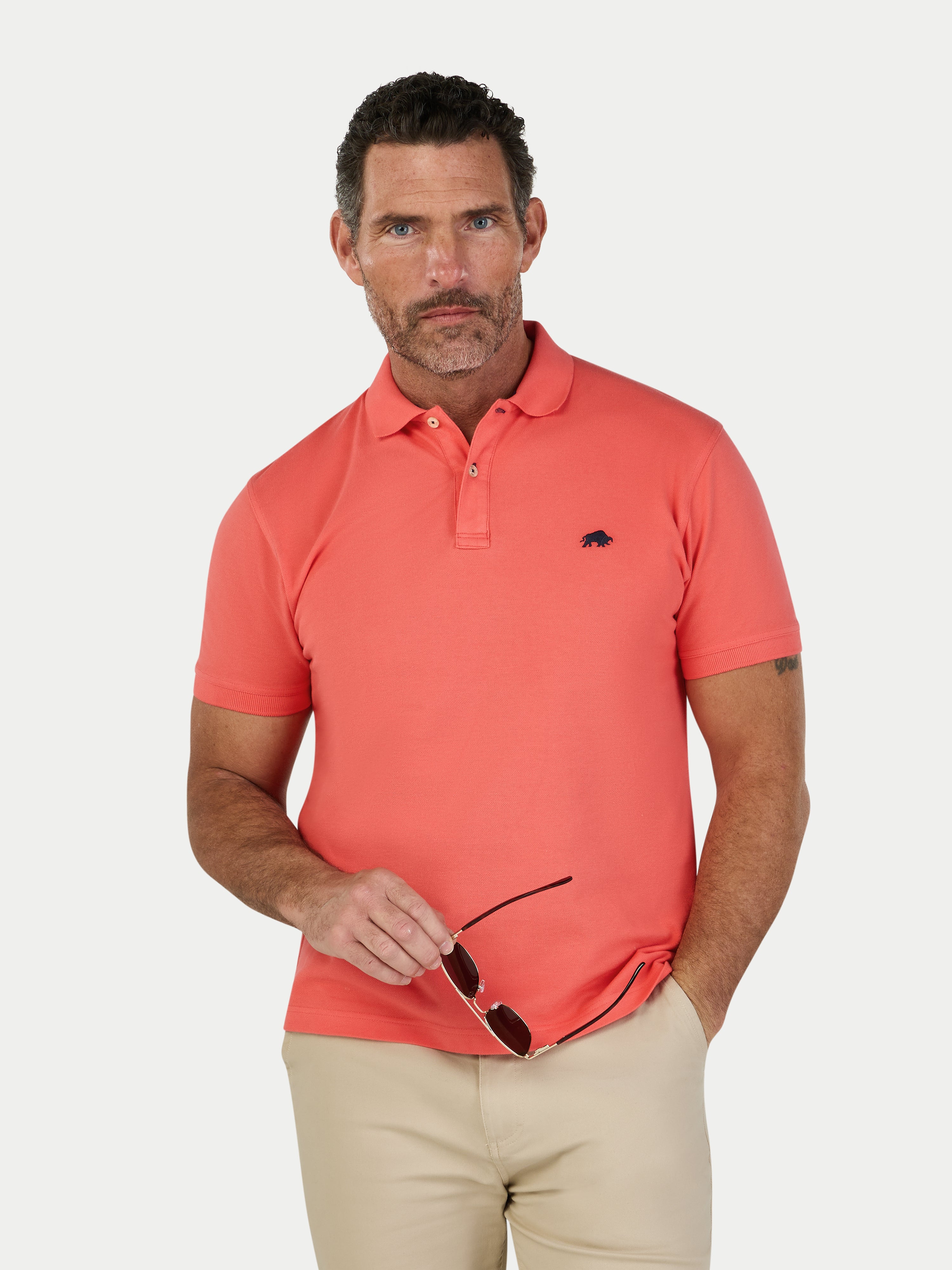 Man wearing a coral polo shirt with a logo, holding sunglasses, on a white background