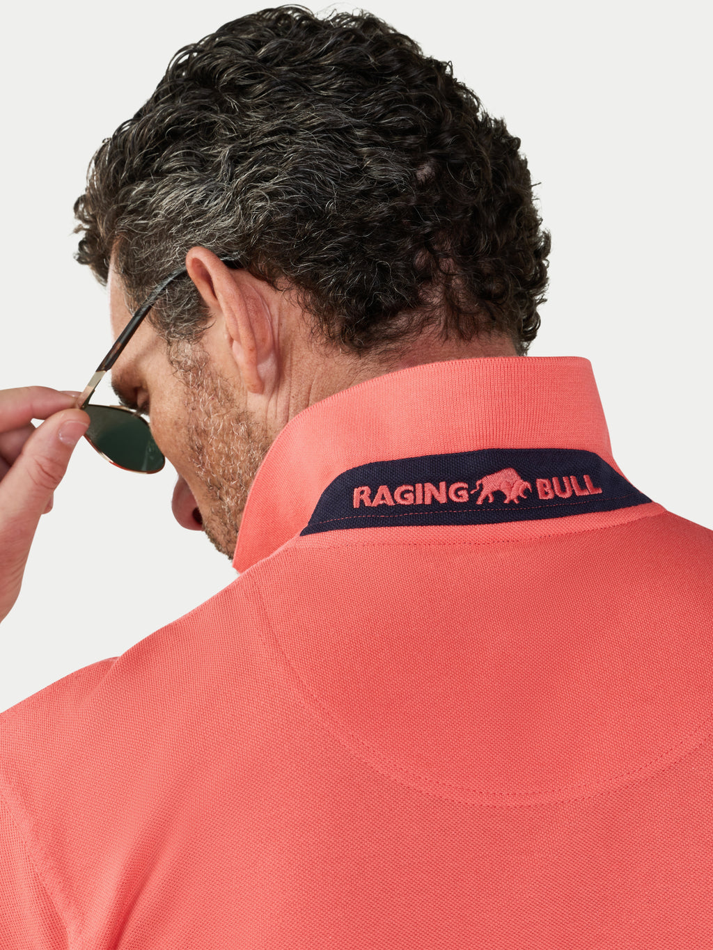 Man wearing a watermelon coloured polo shirt with 'Raging Bull' logo, looking over sunglasses.