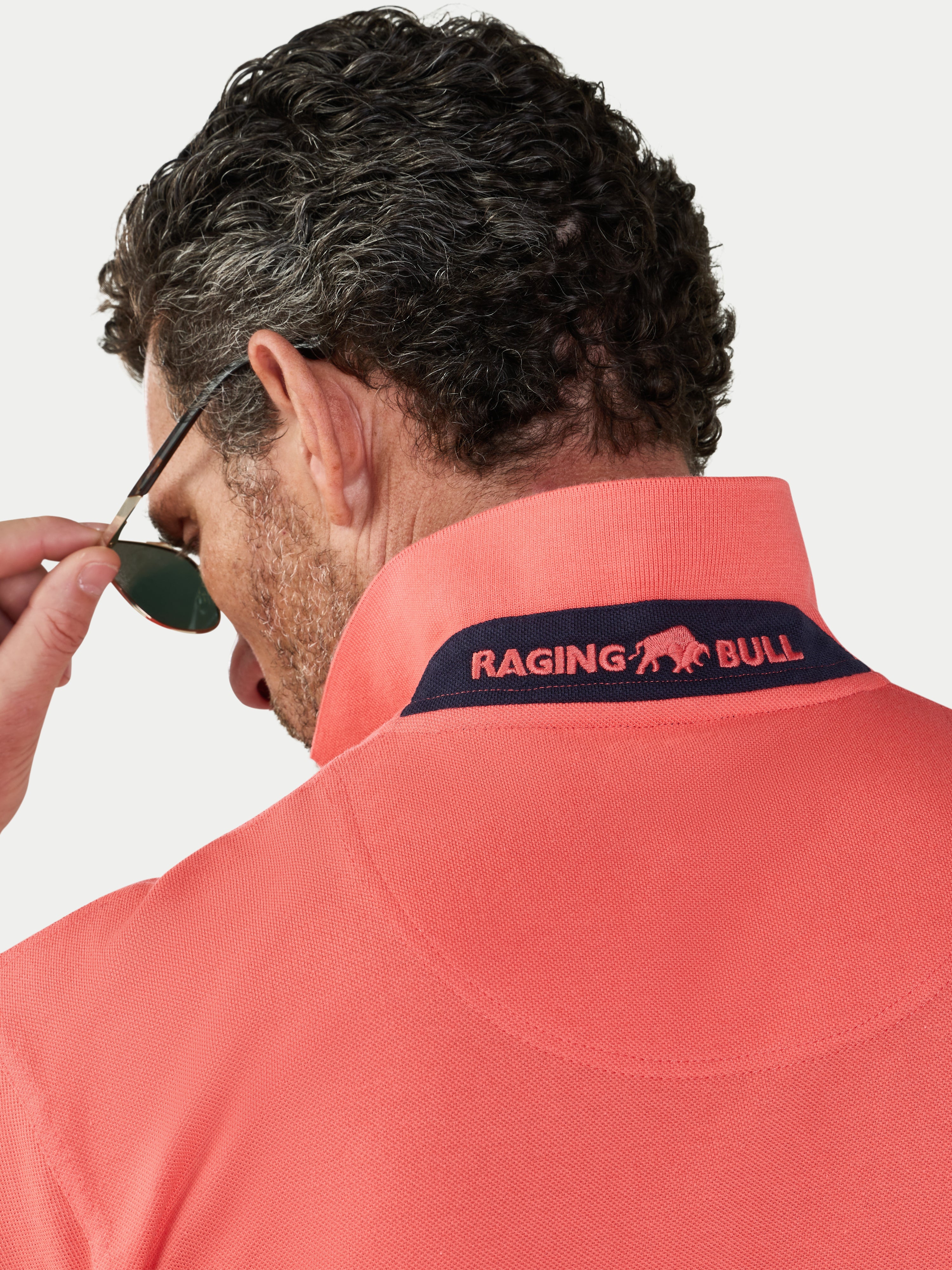 Man wearing a watermelon coloured polo shirt with 'Raging Bull' logo, looking over sunglasses.