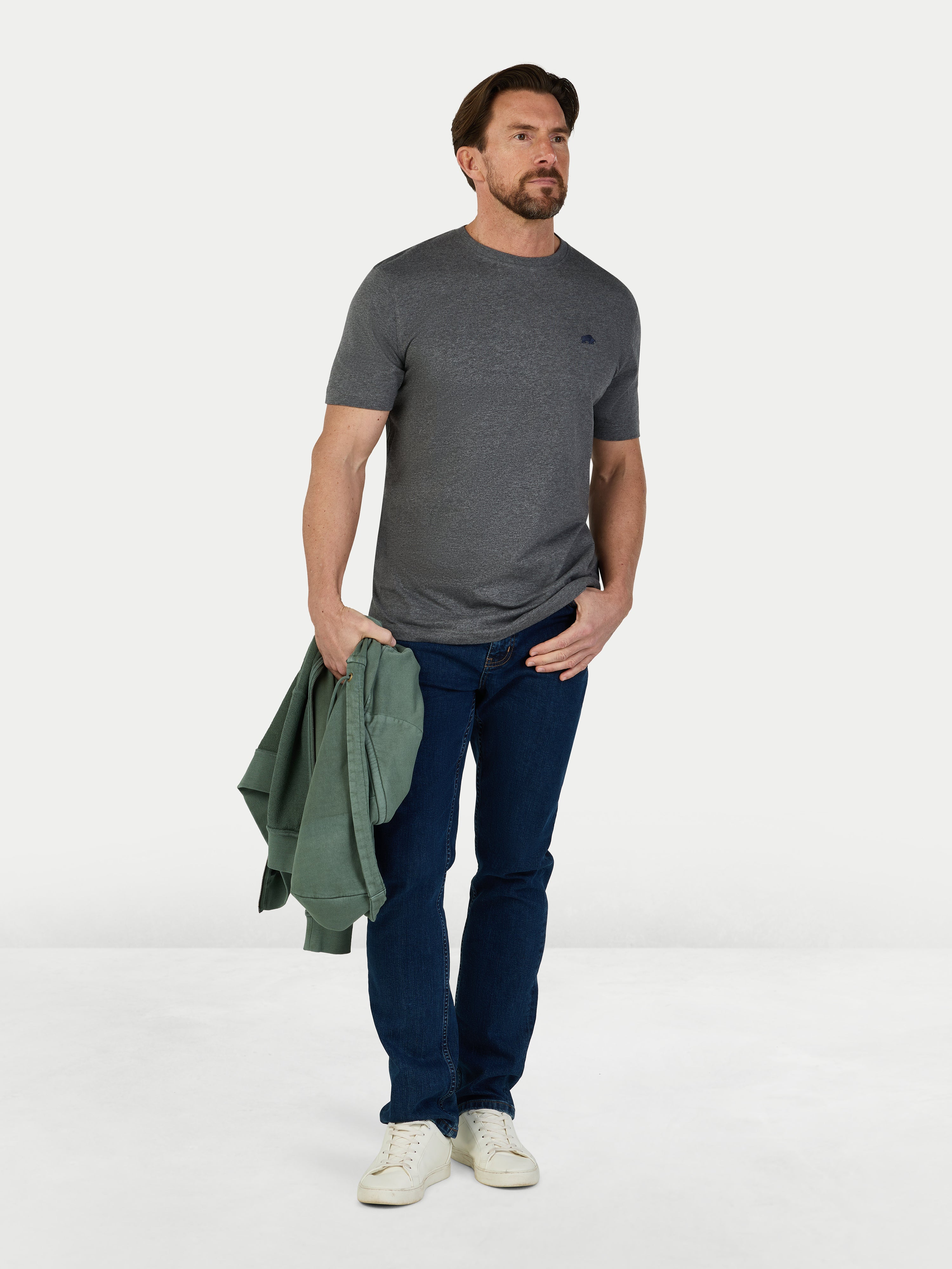 Man wearing a gray t-shirt, blue jeans, and white sneakers holding a green jacket on a white background