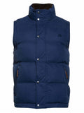 A male model wearing a navy padded gilet with zip pockets and a zip through fastening, standing in front of a sports stadium seating.