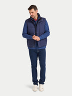 Man wearing a blue vest over a blue shirt with dark jeans on a white background