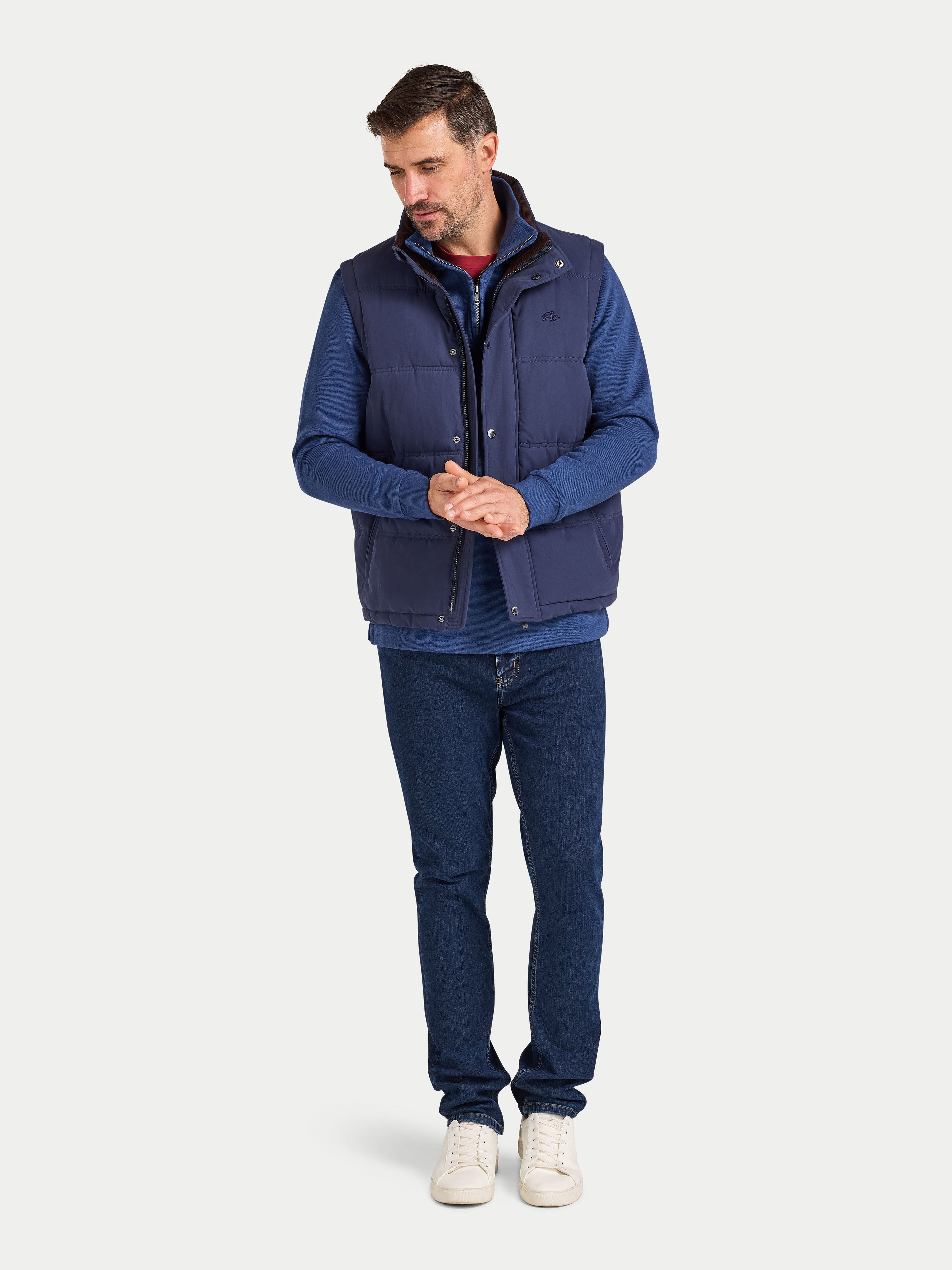 Man wearing a blue vest over a blue shirt with dark jeans on a white background