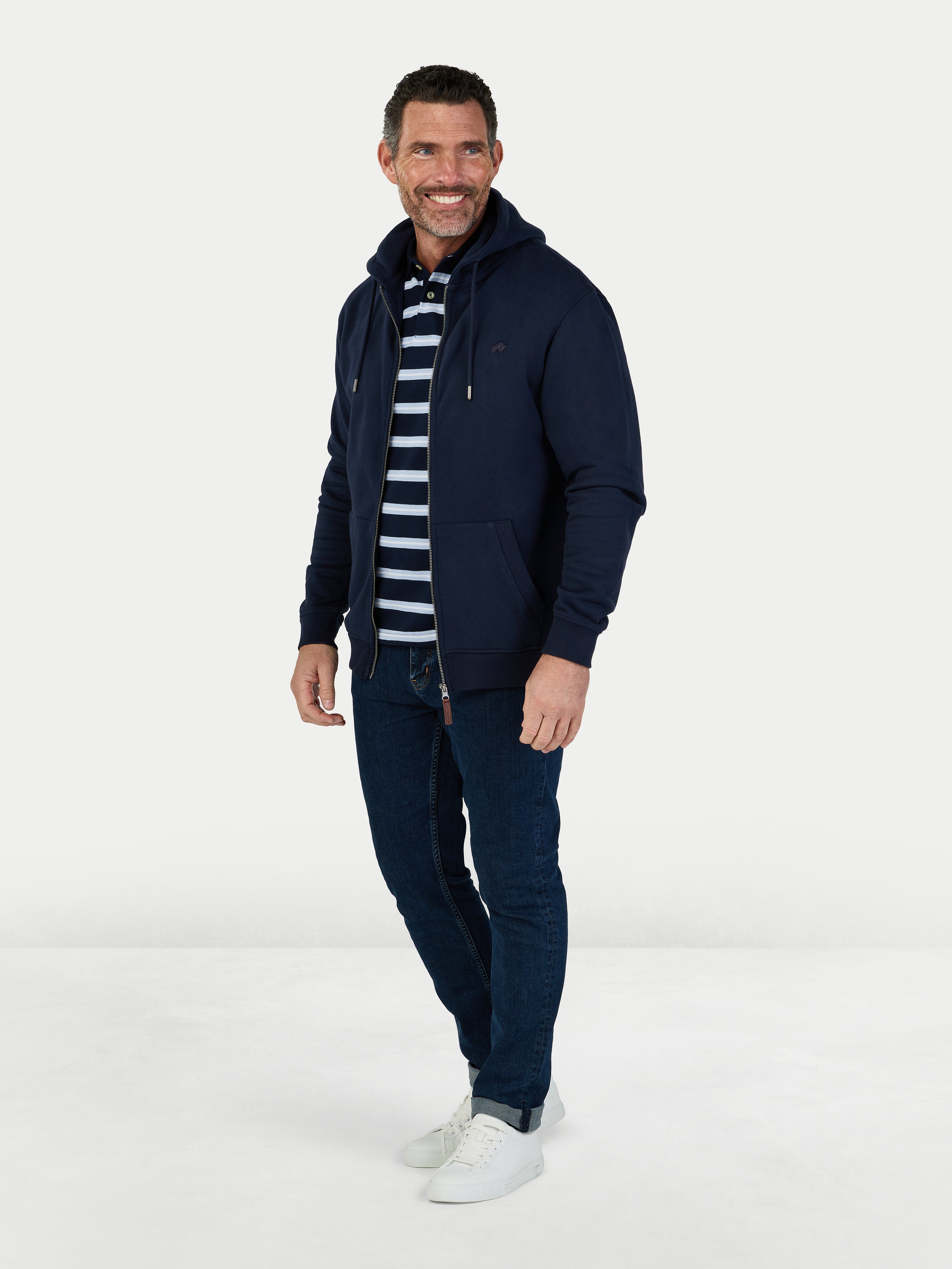 Man wearing a navy blue jacket, striped shirt, and jeans on a white background
