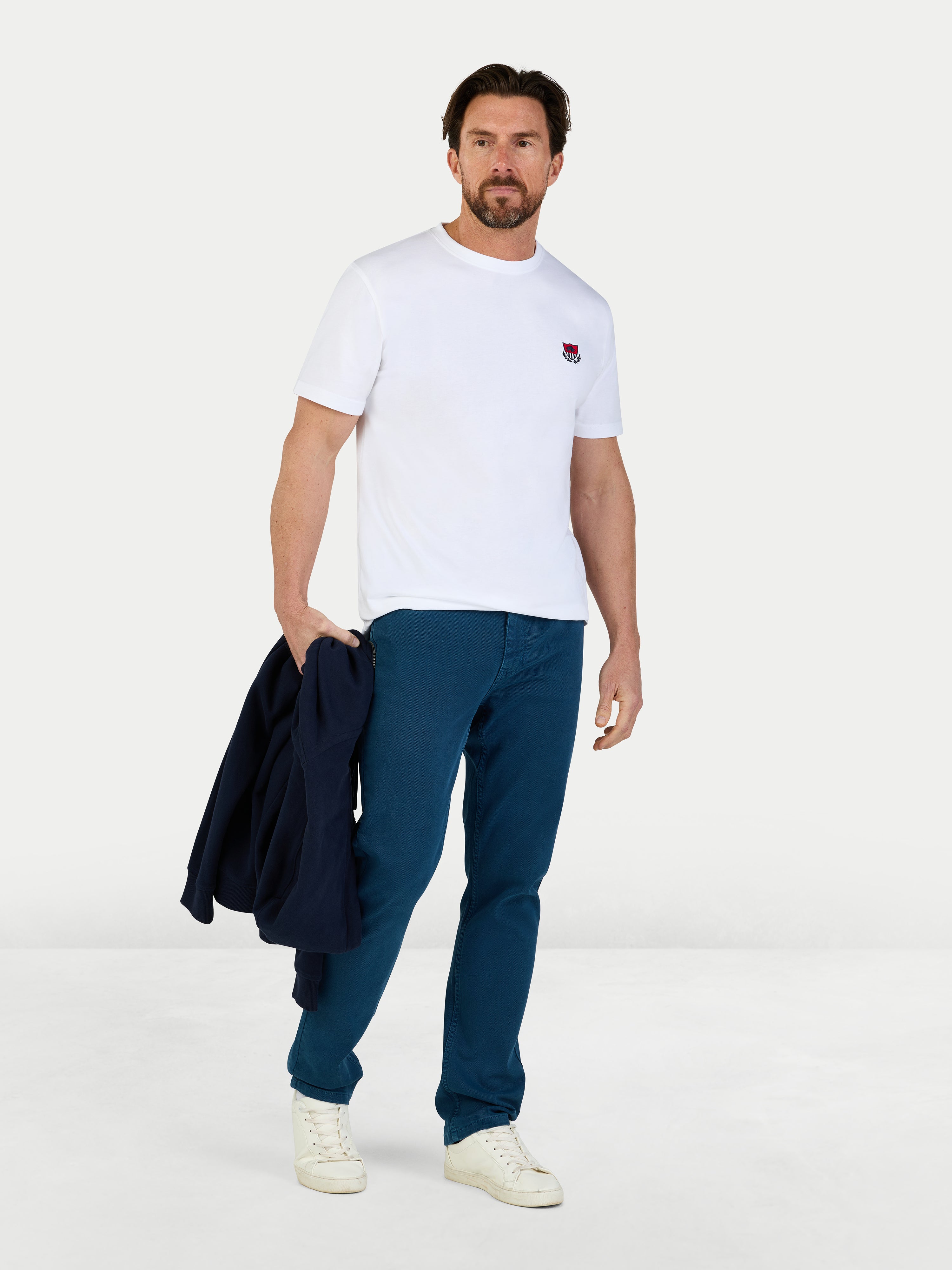 Man wearing a white t-shirt and blue pants holding a jacket on a white background