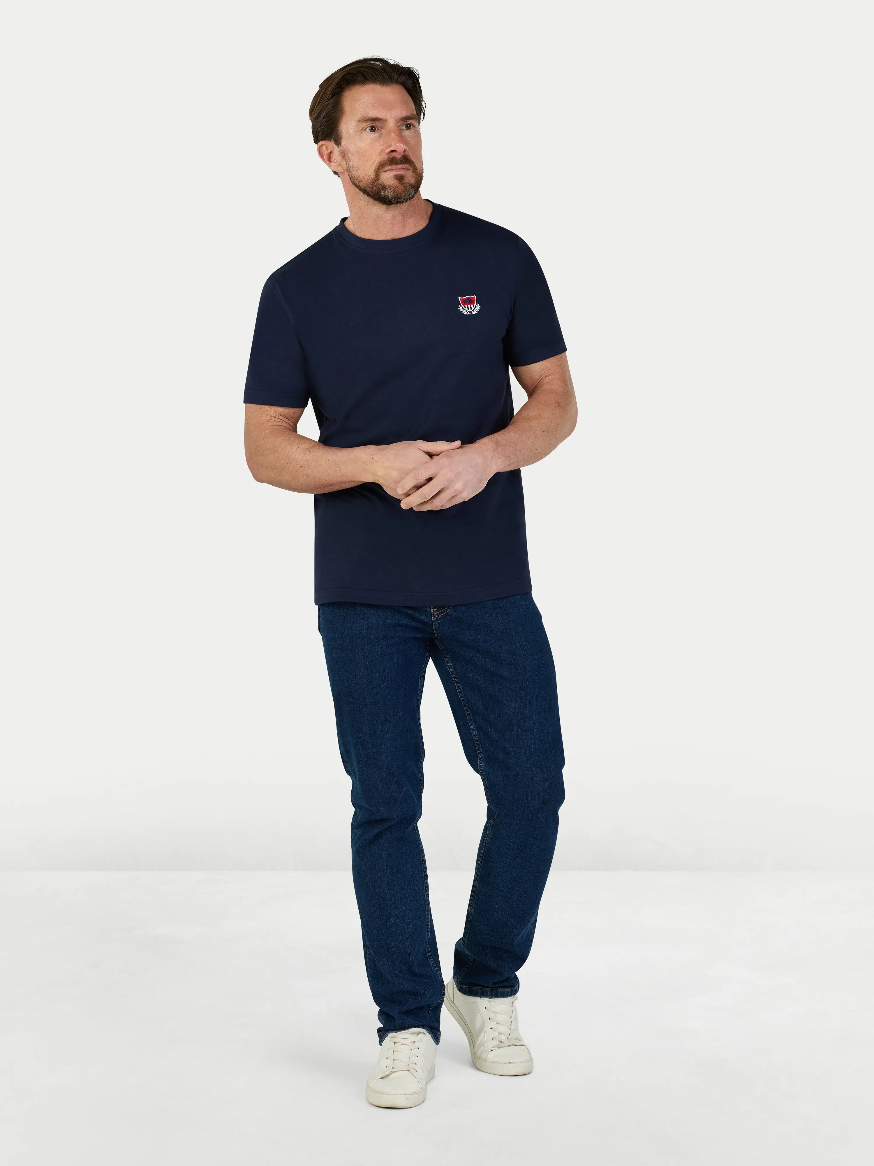 Man wearing a navy t-shirt with a logo and dark jeans on a white background