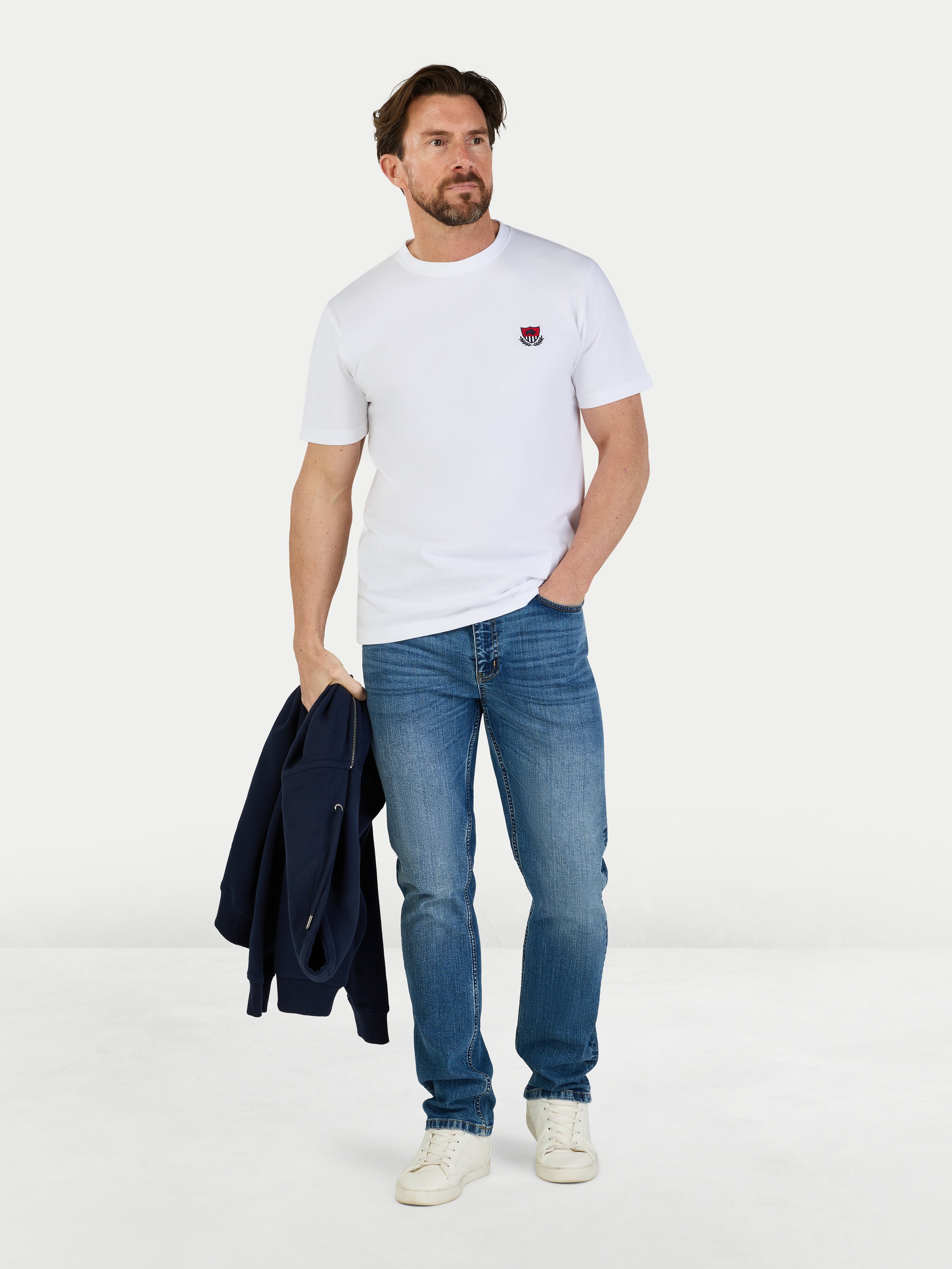 Man wearing a white t-shirt, blue jeans, and white sneakers holding a dark jacket on a light gray background