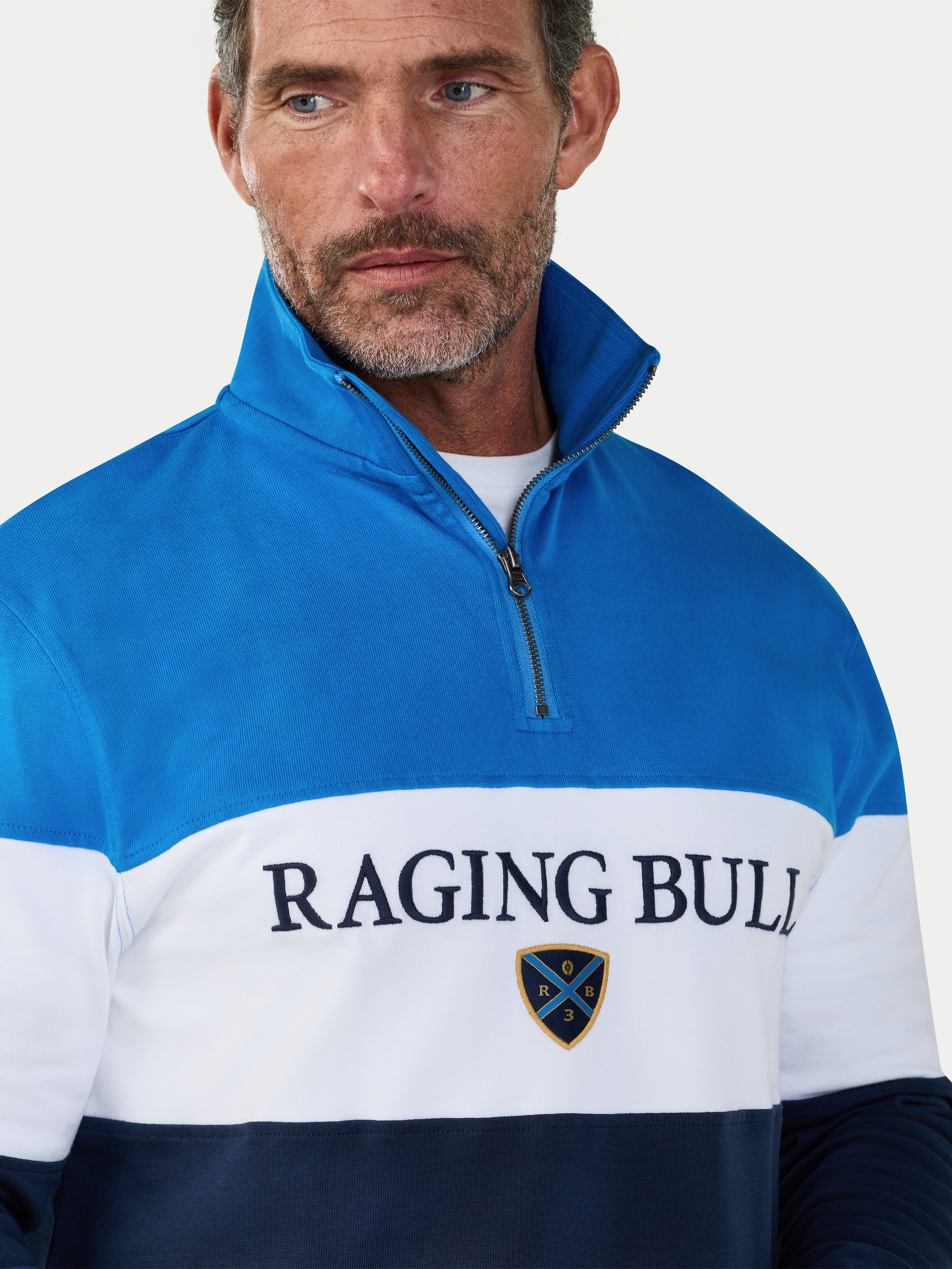 Man wearing a blue and white pullover with 'Raging Bull' branding on a light gray background