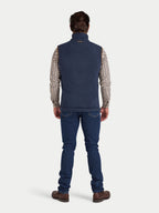 Man wearing a navy blue vest over a plaid shirt with blue jeans on a white background