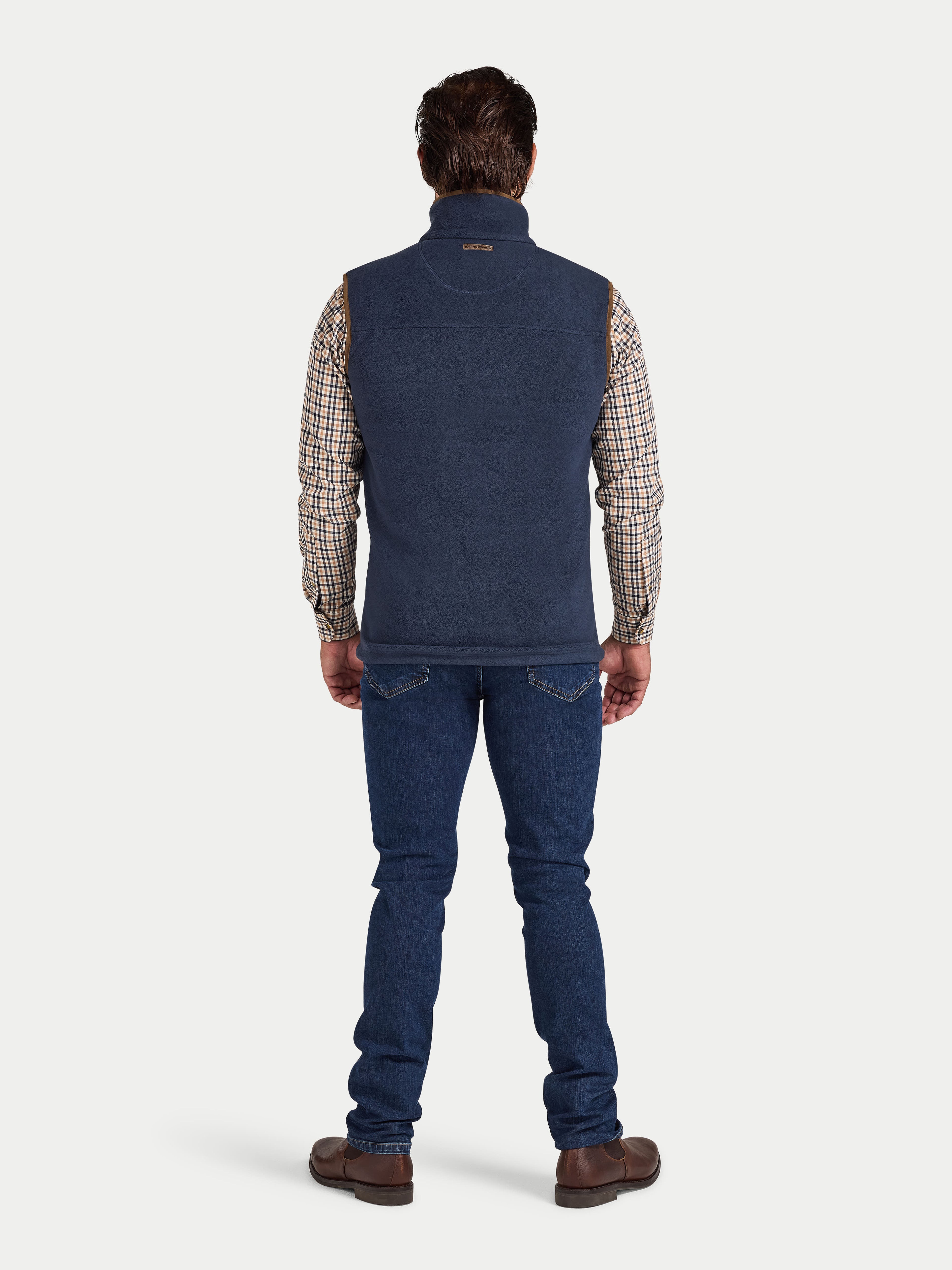 Man wearing a navy blue vest over a plaid shirt with blue jeans on a white background