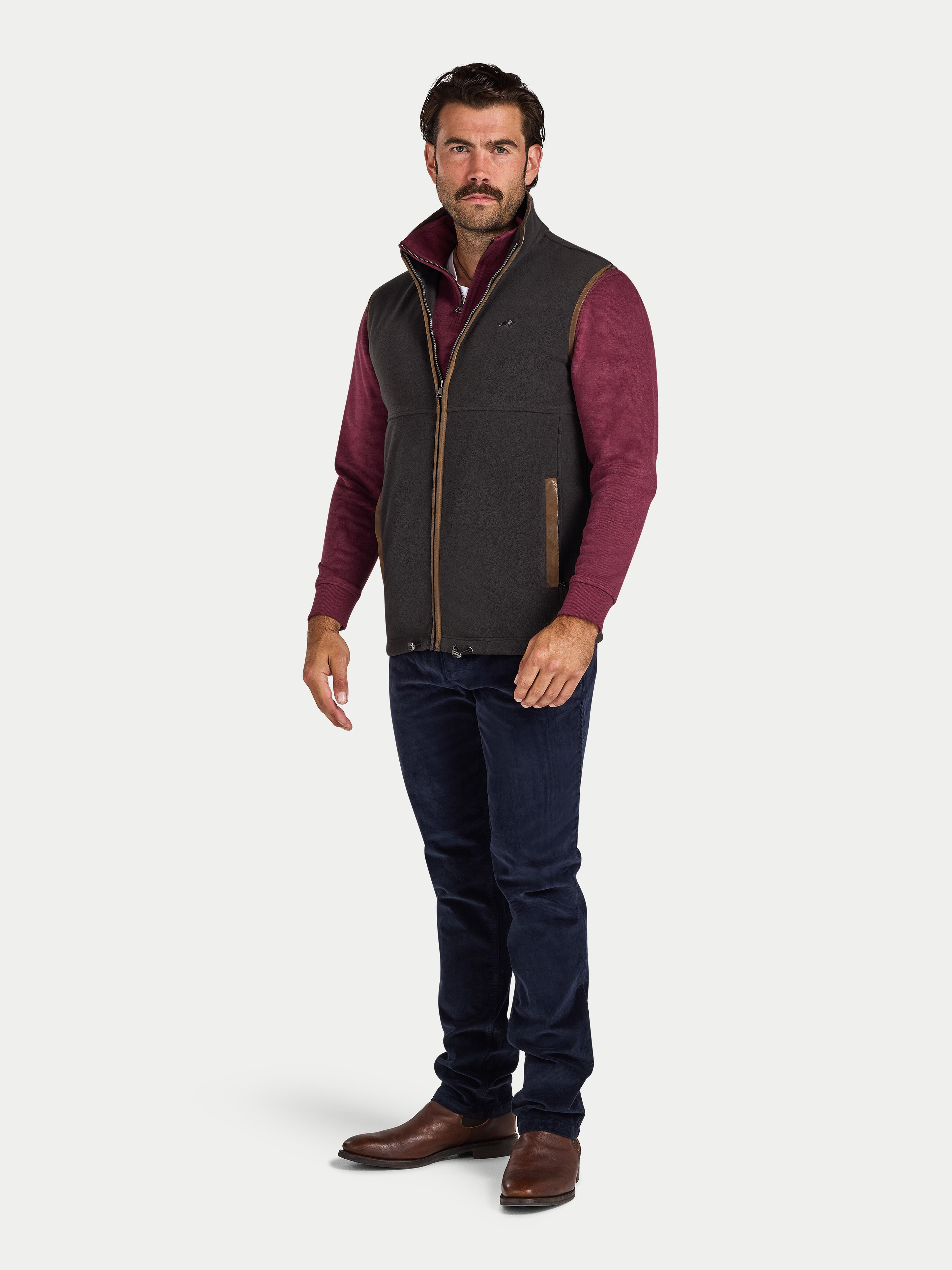 Man wearing a slate coloured gilet over a red shirt with navy pants on a white background