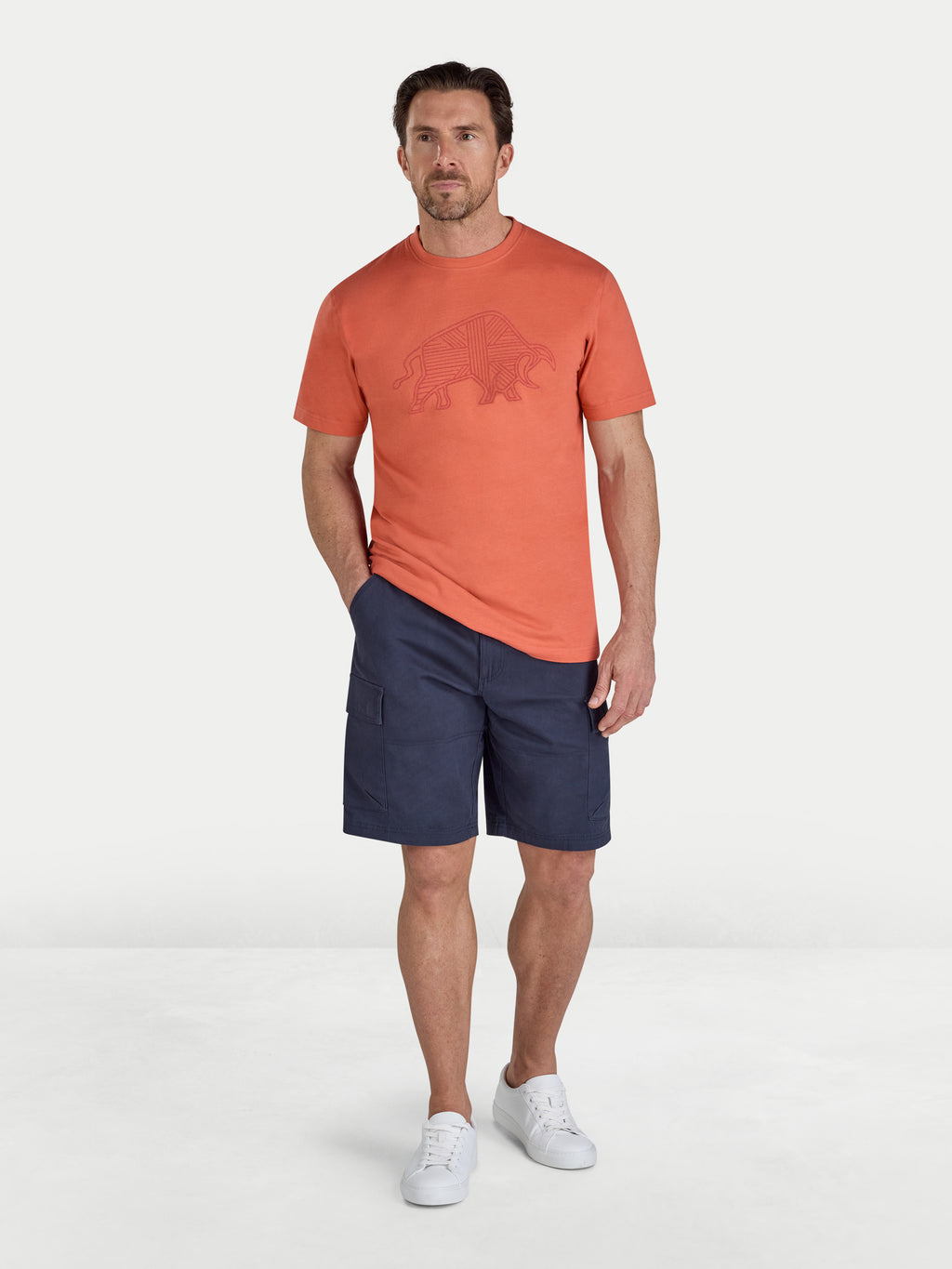 Man wearing an orange t-shirt with a logo and navy shorts on a white background