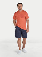Man wearing an orange t-shirt with a logo and navy shorts on a white background