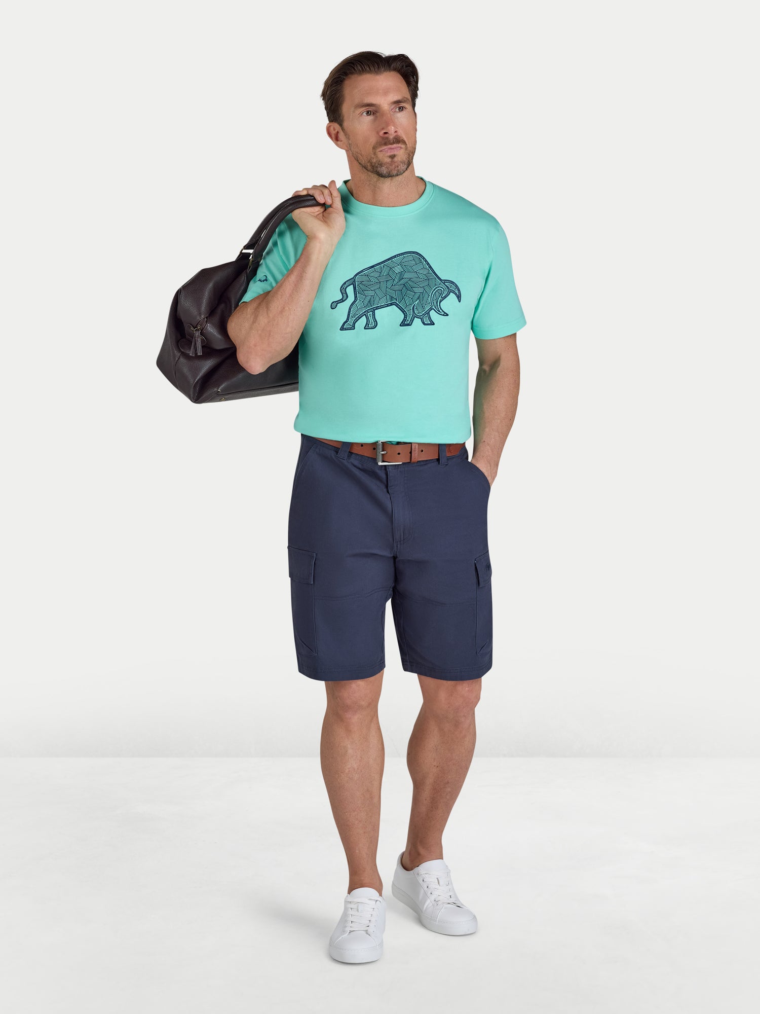 Man wearing a mint green t-shirt with a graphic design and navy shorts, holding a black bag on a white background.