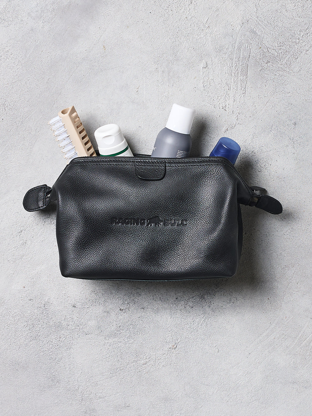 Black leather toiletry bag with travel items on a gray surface