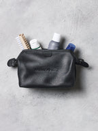 Black leather toiletry bag with travel items on a gray surface
