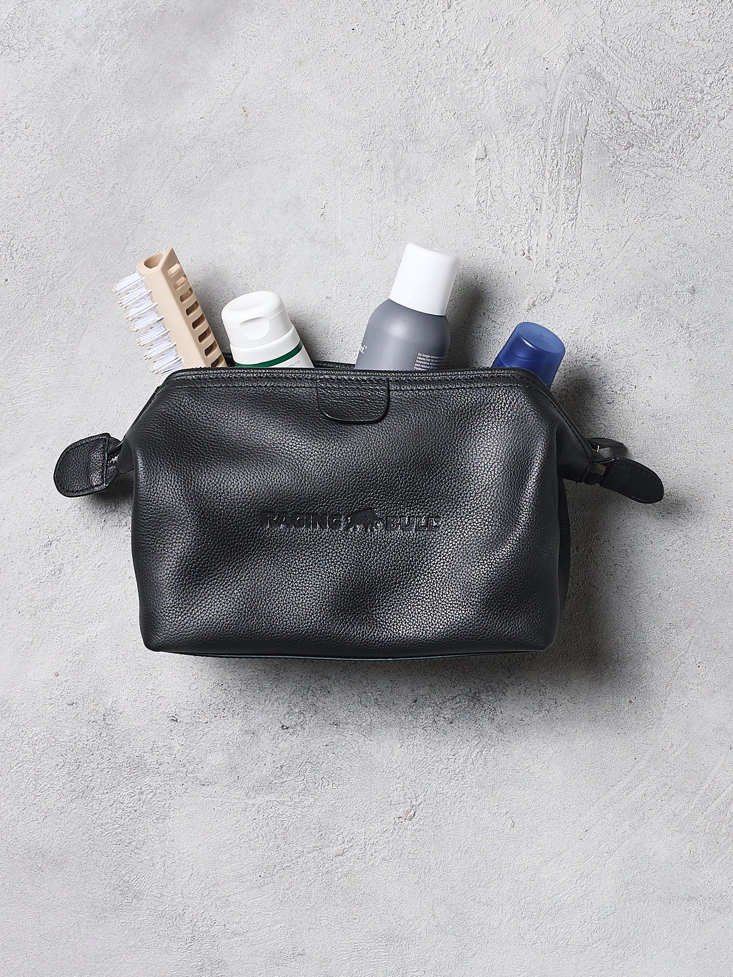Black leather toiletry bag with travel items on a gray surface