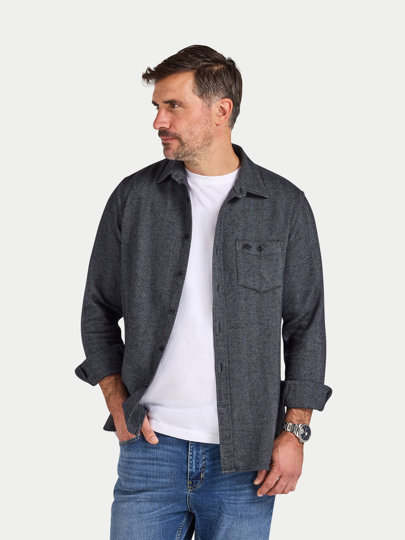 Man wearing a dark denim shirt over a white t-shirt with blue jeans on a light gray background