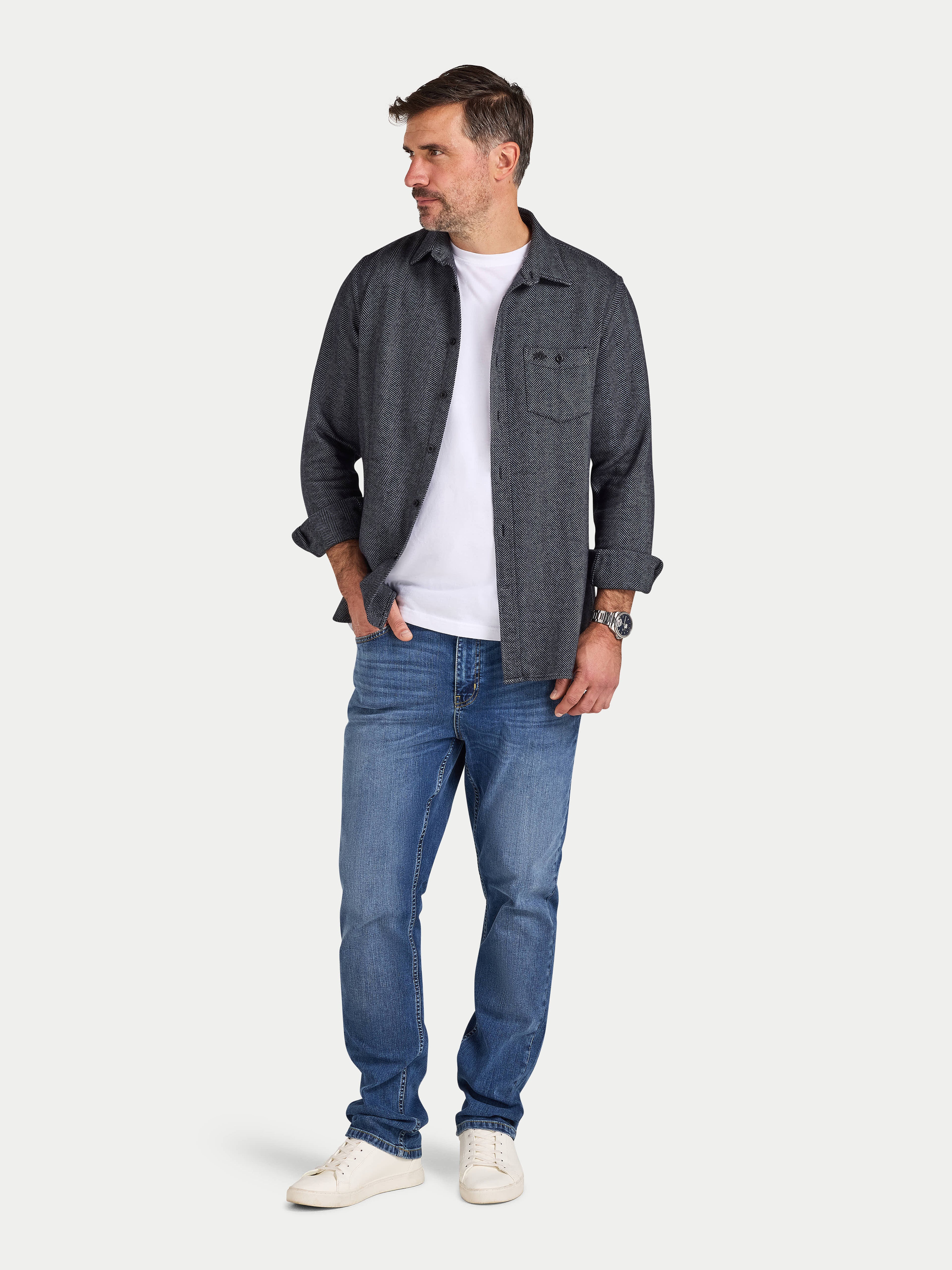 Man wearing a dark denim shirt over a white t-shirt, paired with blue jeans on a light gray background