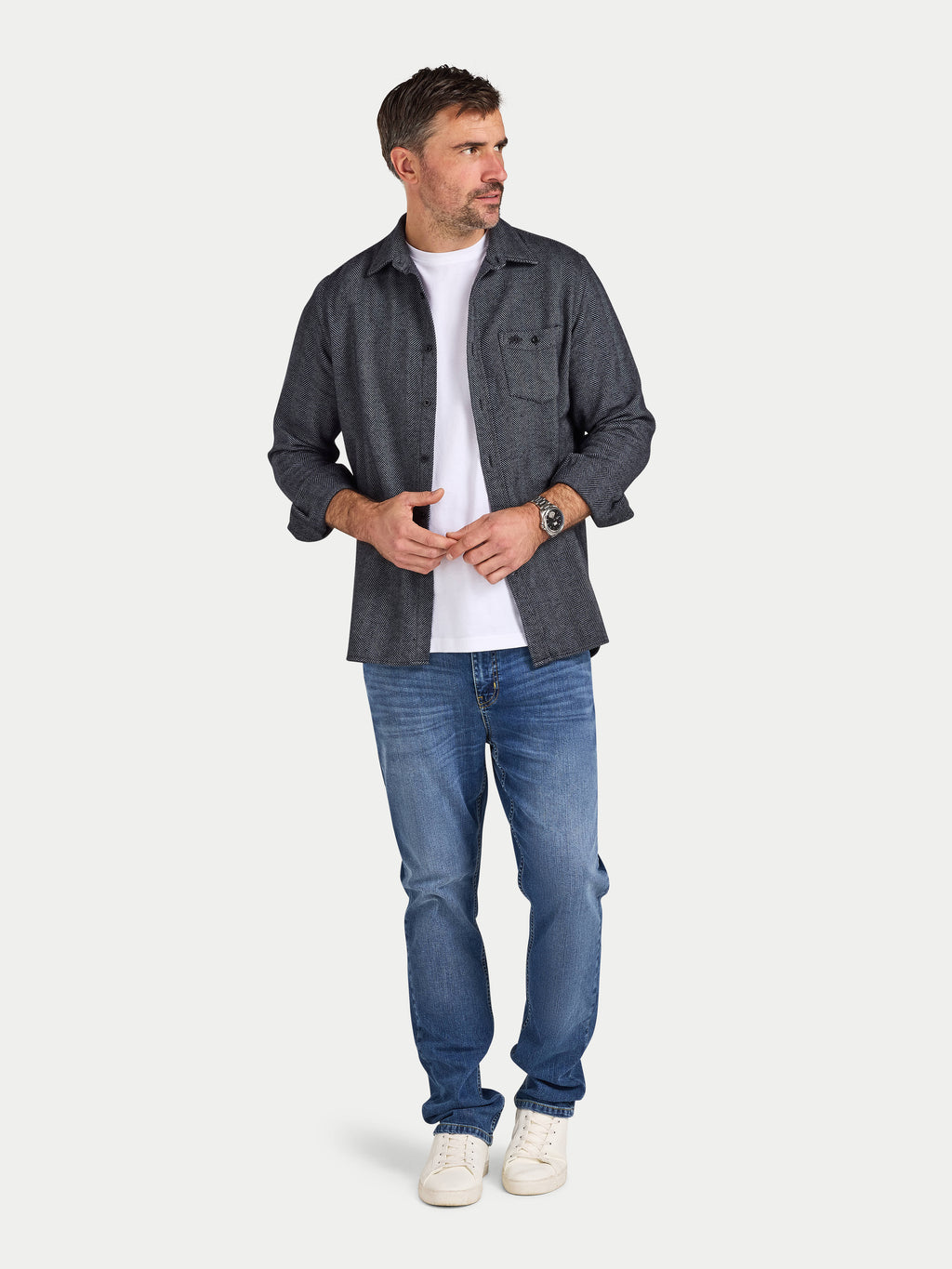 Man wearing a dark gray flannel shirt, white t-shirt, blue jeans, and white sneakers on a light gray background