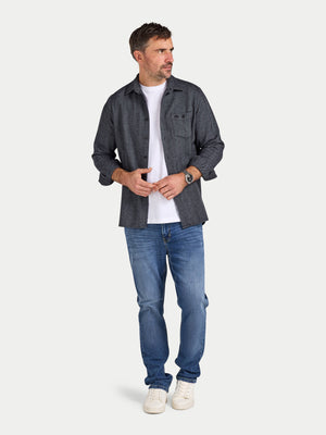 Man wearing a dark gray flannel shirt, white t-shirt, blue jeans, and white sneakers on a light gray background