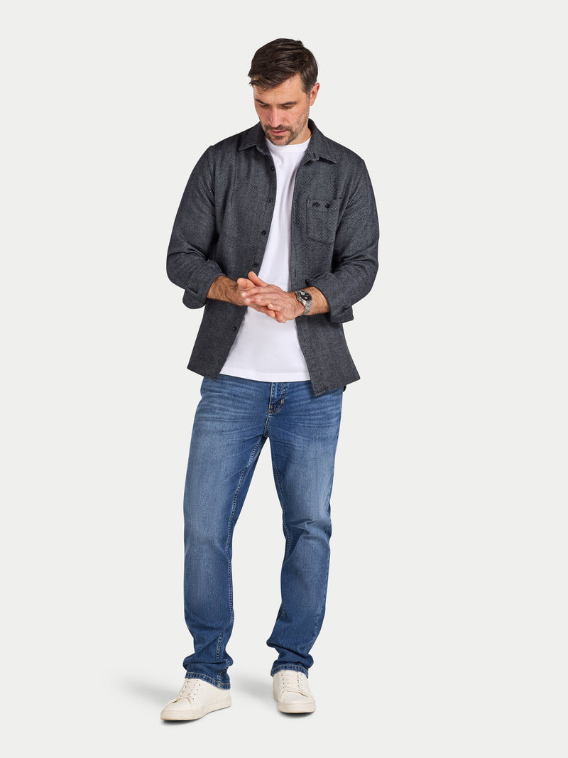 Man wearing a dark gray flannel shirt, white t-shirt, blue jeans, and white sneakers on a light gray background