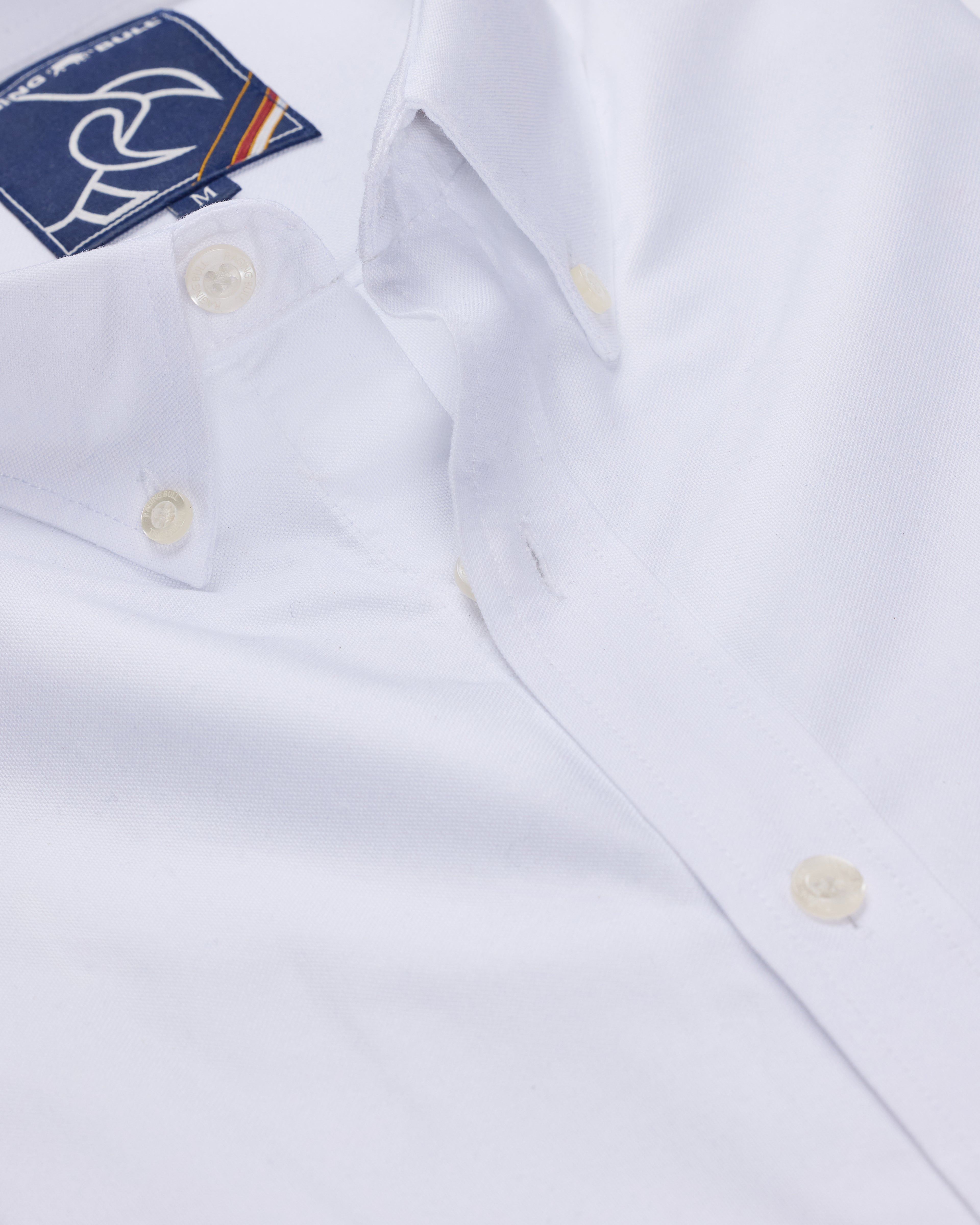 A long sleeve classic Oxford shirt in white, showing the collar, button-up design, and a small brand tag on the upper chest.