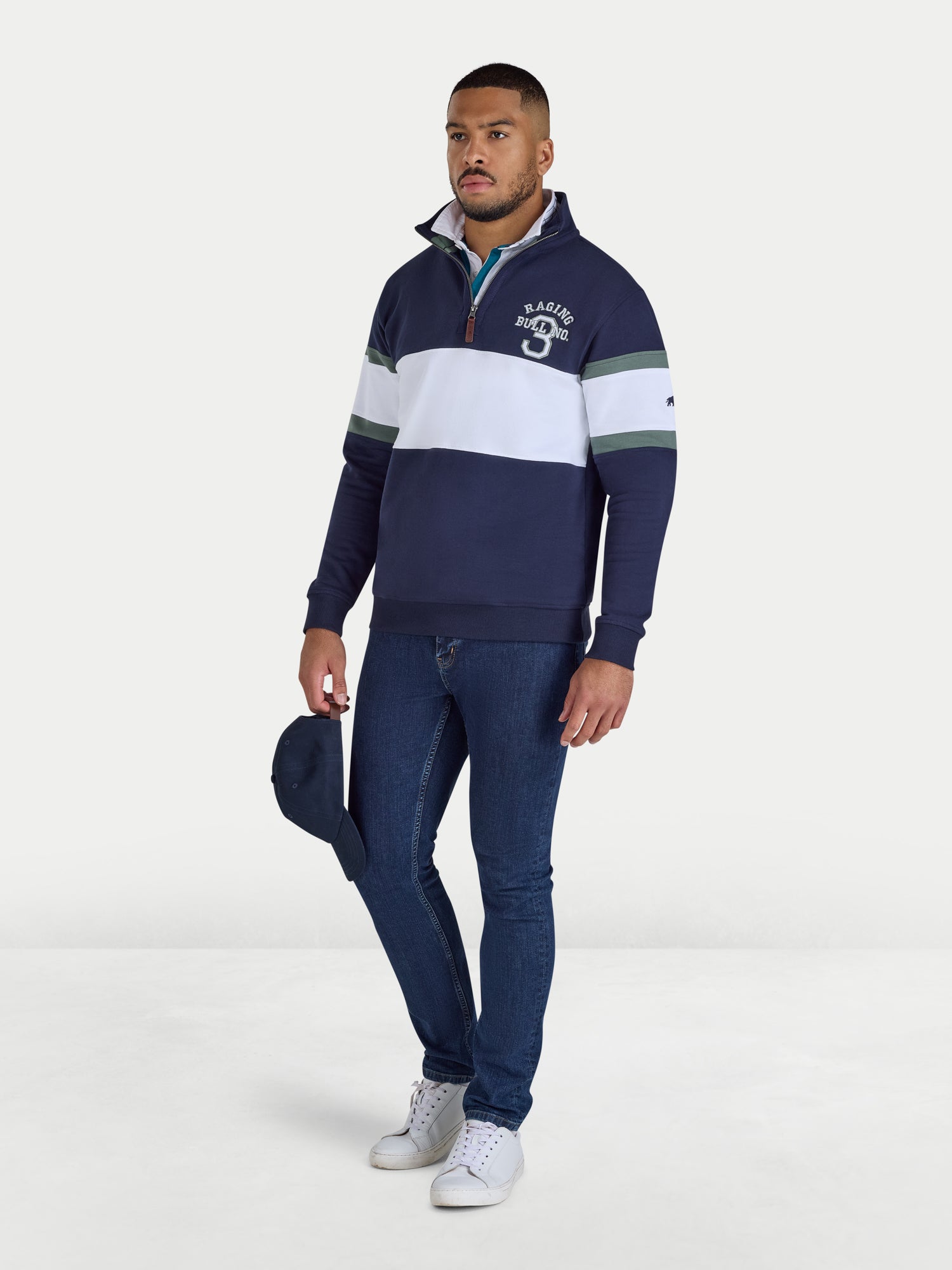 Man wearing a navy and white striped sweater with a logo, holding a blue cap on a white background