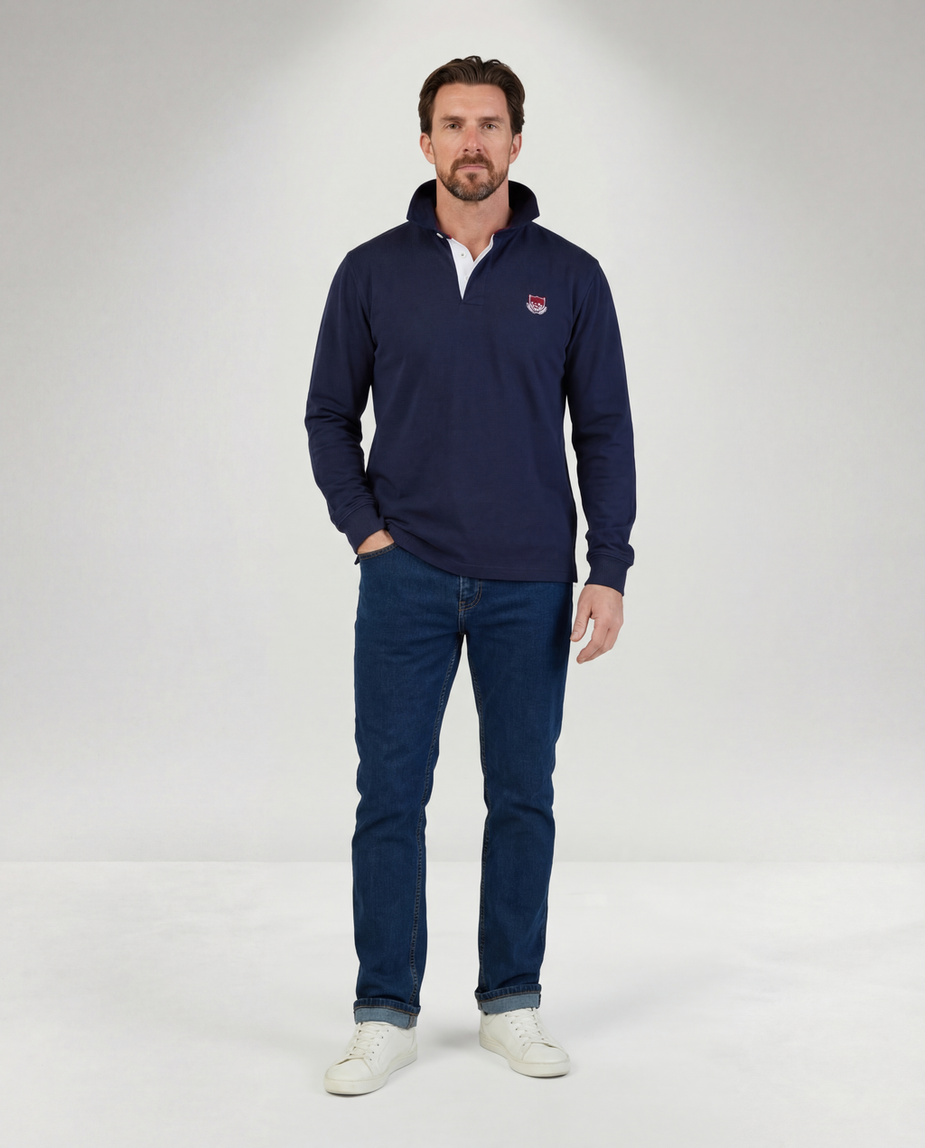 Man wearing a navy long-sleeve shirt with a logo and blue jeans on a white background