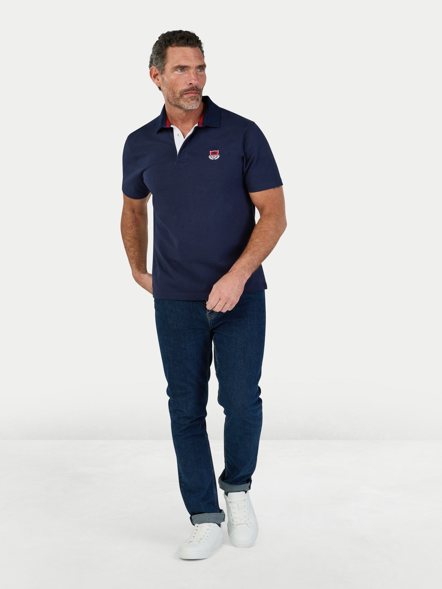 Man wearing a navy polo shirt with a logo and dark jeans on a white background
