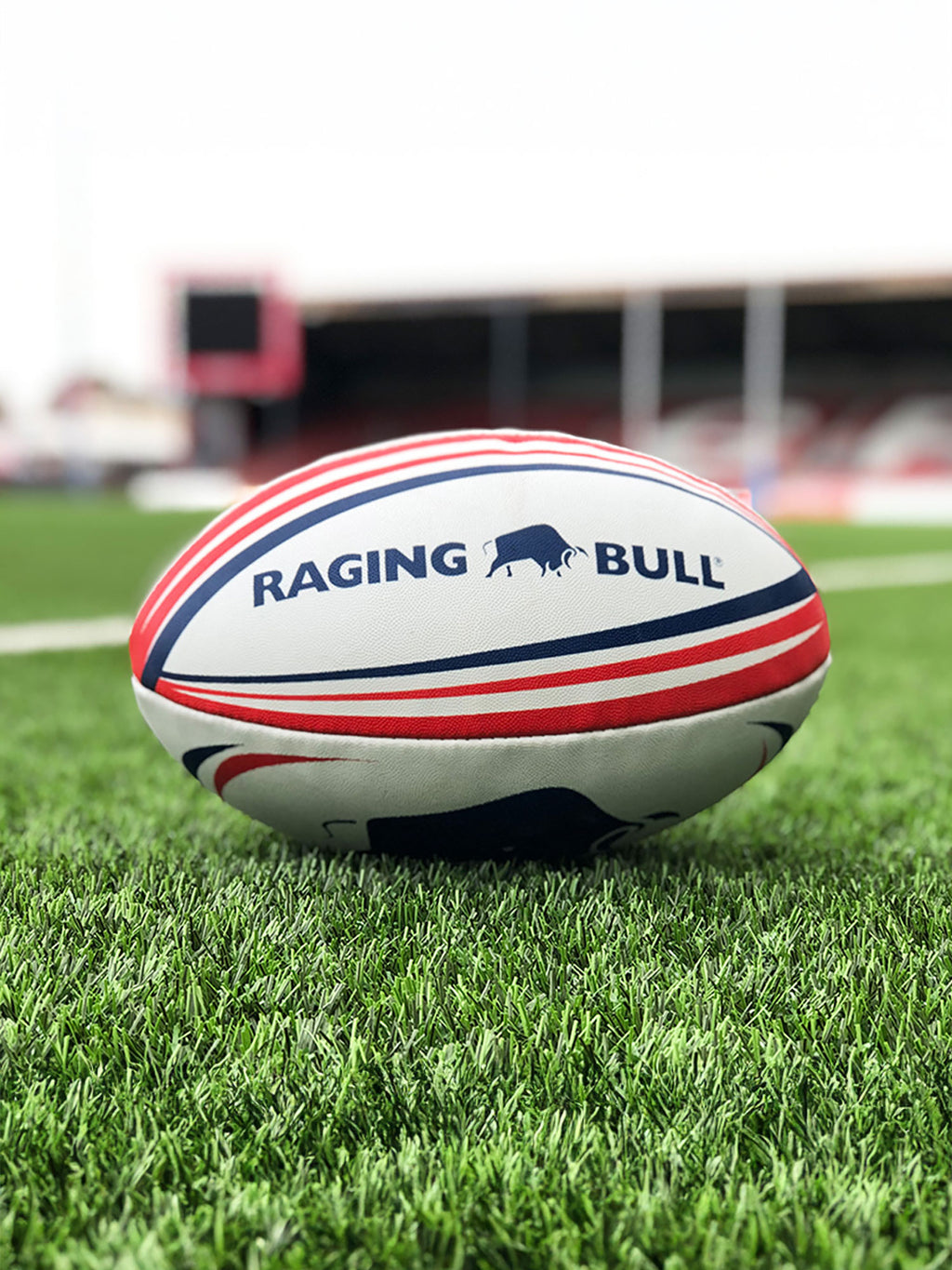A white rugby ball with red and blue accents lying on grass, featuring the 'Raging Bull' brand logo.