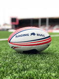 A white rugby ball with red and blue accents lying on grass, featuring the 'Raging Bull' brand logo.