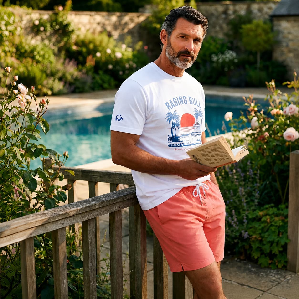 Man wearing Raging Bull beach rygby t-shirt in white and swimming shorts in coral with a pool and a beautiful English garden in the background.