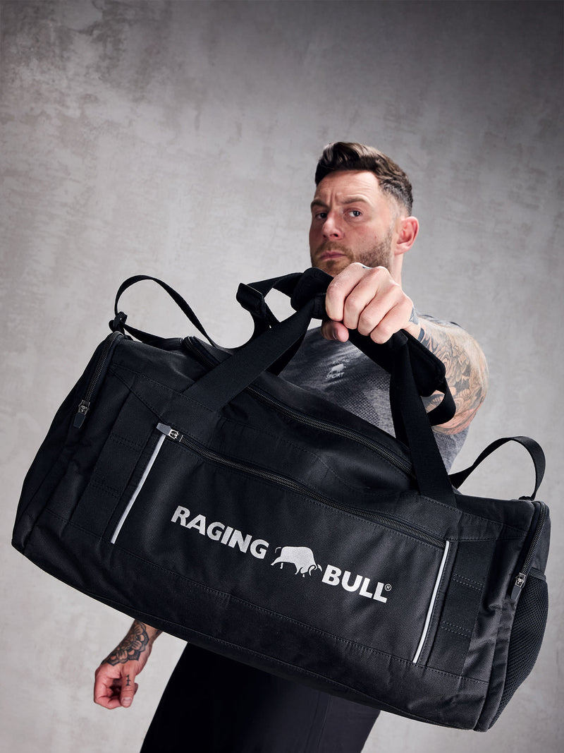 A man holding a black duffle bag with 'RAGING BULL' text on it.