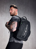 A man wearing a black rucksack with adjustable shoulder straps and a padded mesh back. The backpack features multiple compartments, including a zipped front compartment and a shoe compartment.