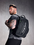 A man wearing a black rucksack with adjustable shoulder straps and a padded mesh back. The backpack features multiple compartments, including a zipped front compartment and a shoe compartment.